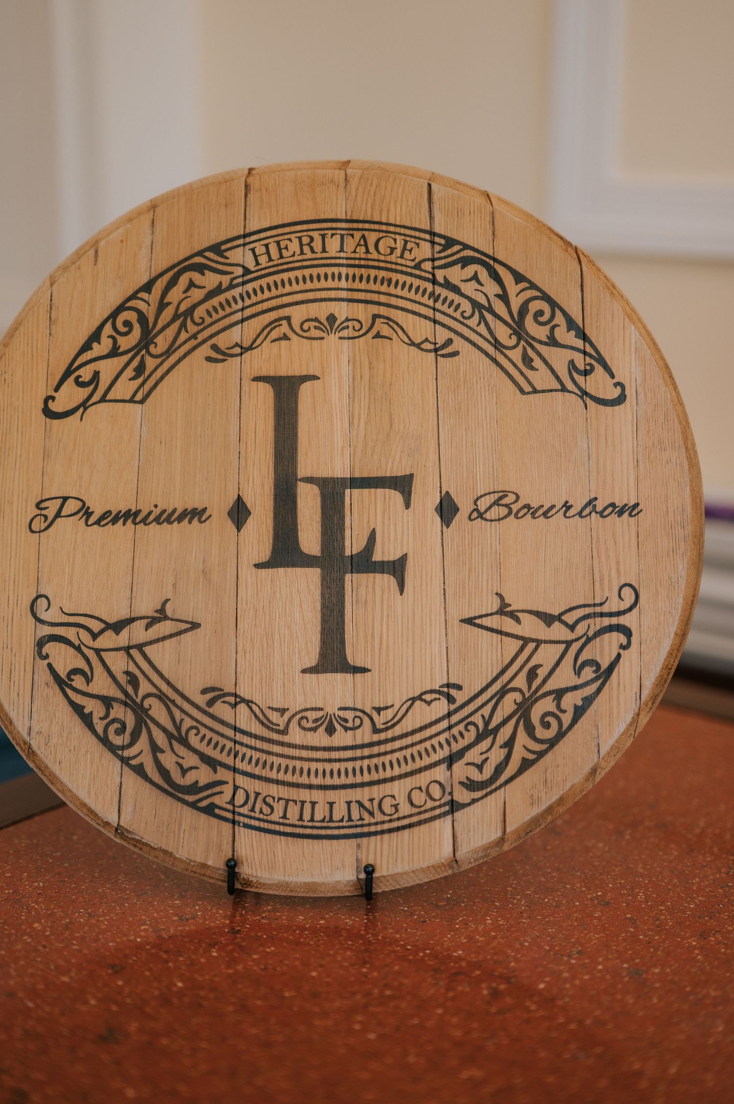 Decorative wooden bourbon barrel lid featuring Heritage Distilling Co.'s LF logo with ornate scrollwork design on natural wood grain.