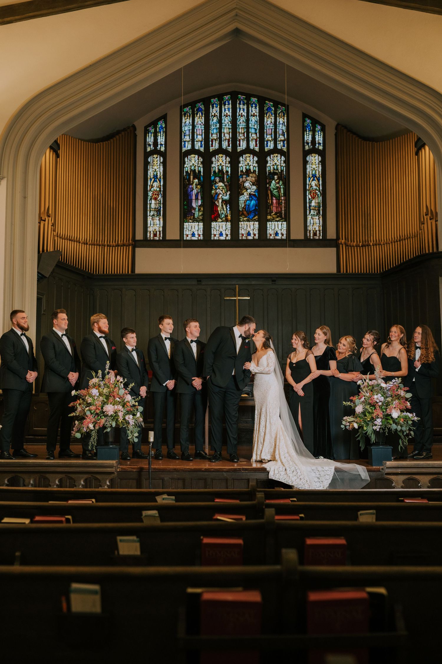 Wedding ceremony in grand church with stained glass windows, organ pipes and bridal party on altar steps.