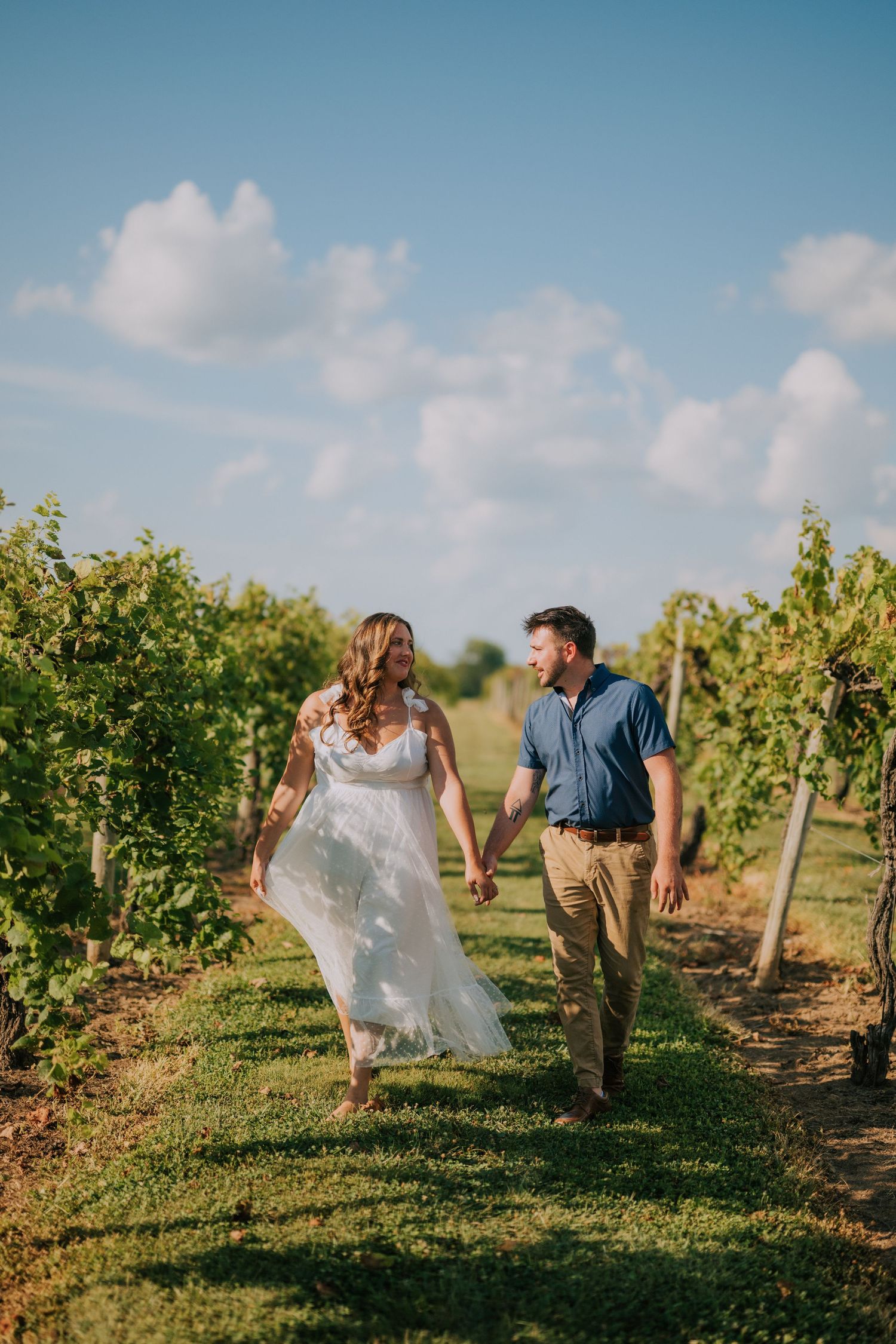 Couple walking through sunny vineyard rows during golden hour photo session.