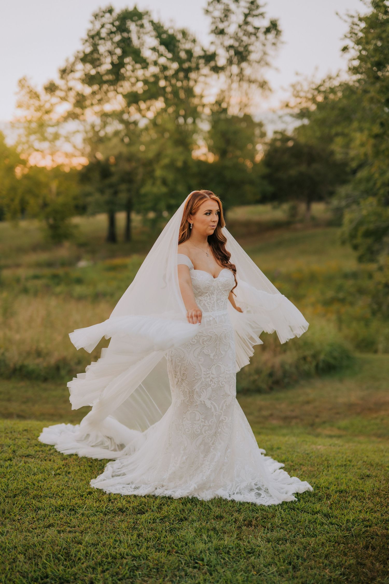 Lace wedding dress spreads beautifully across green hillside during sunset photoshoot.