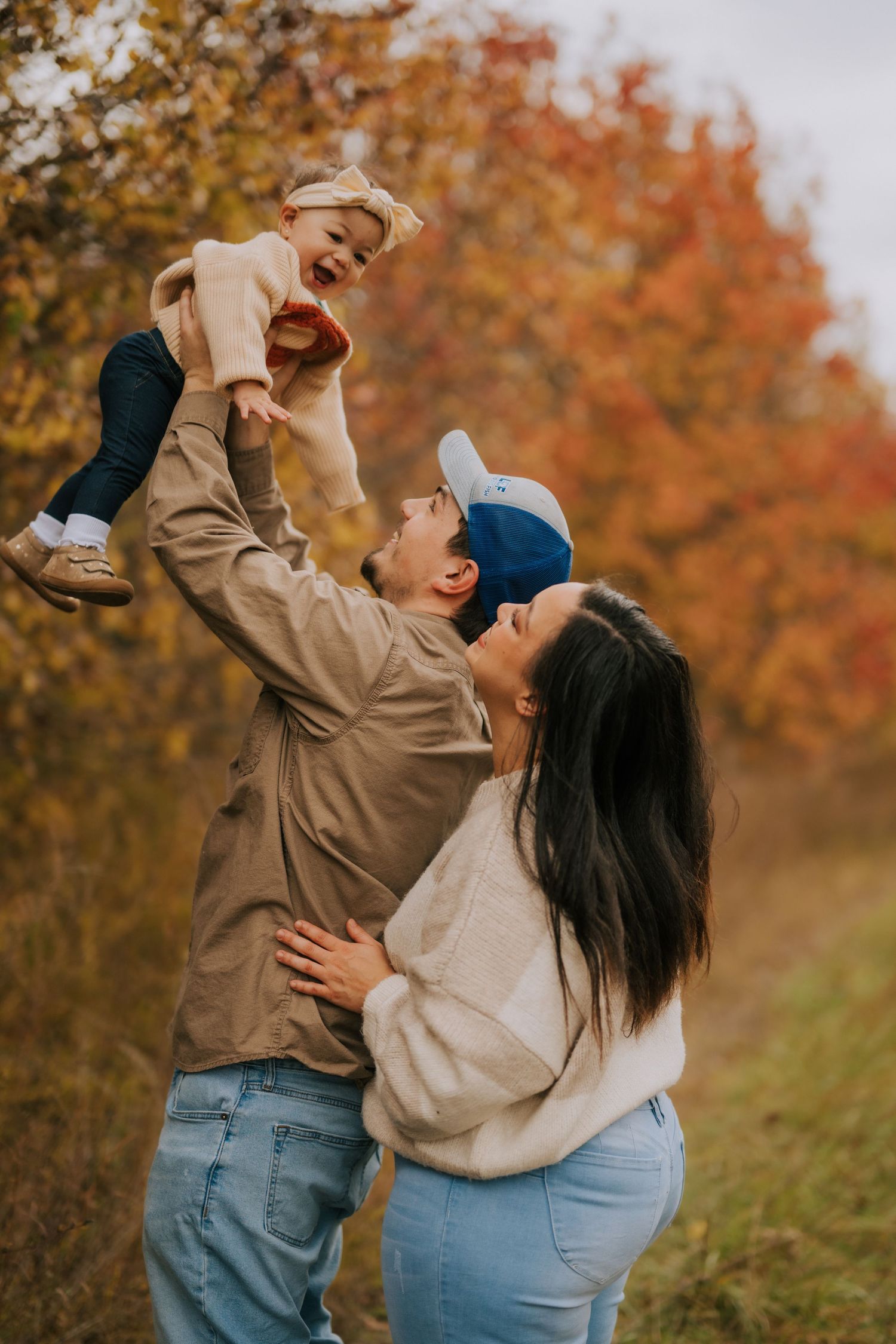 Parents lifting baby in air while playing outdoors among fall foliage.