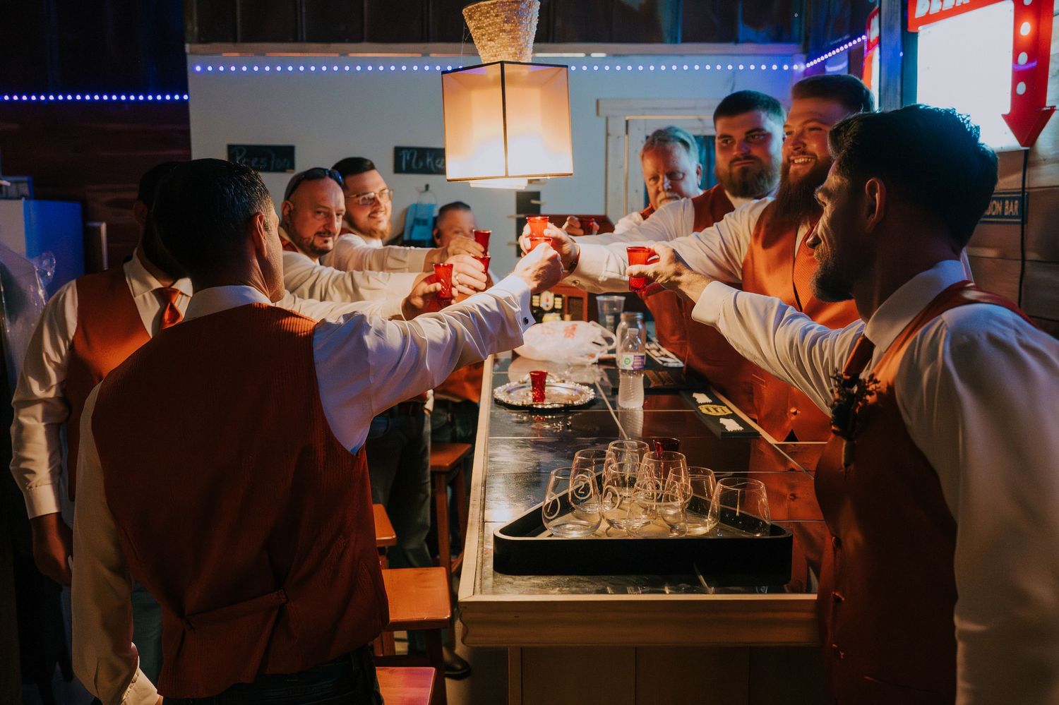 Wedding guests toast and celebrate at an illuminated outdoor bar at night.