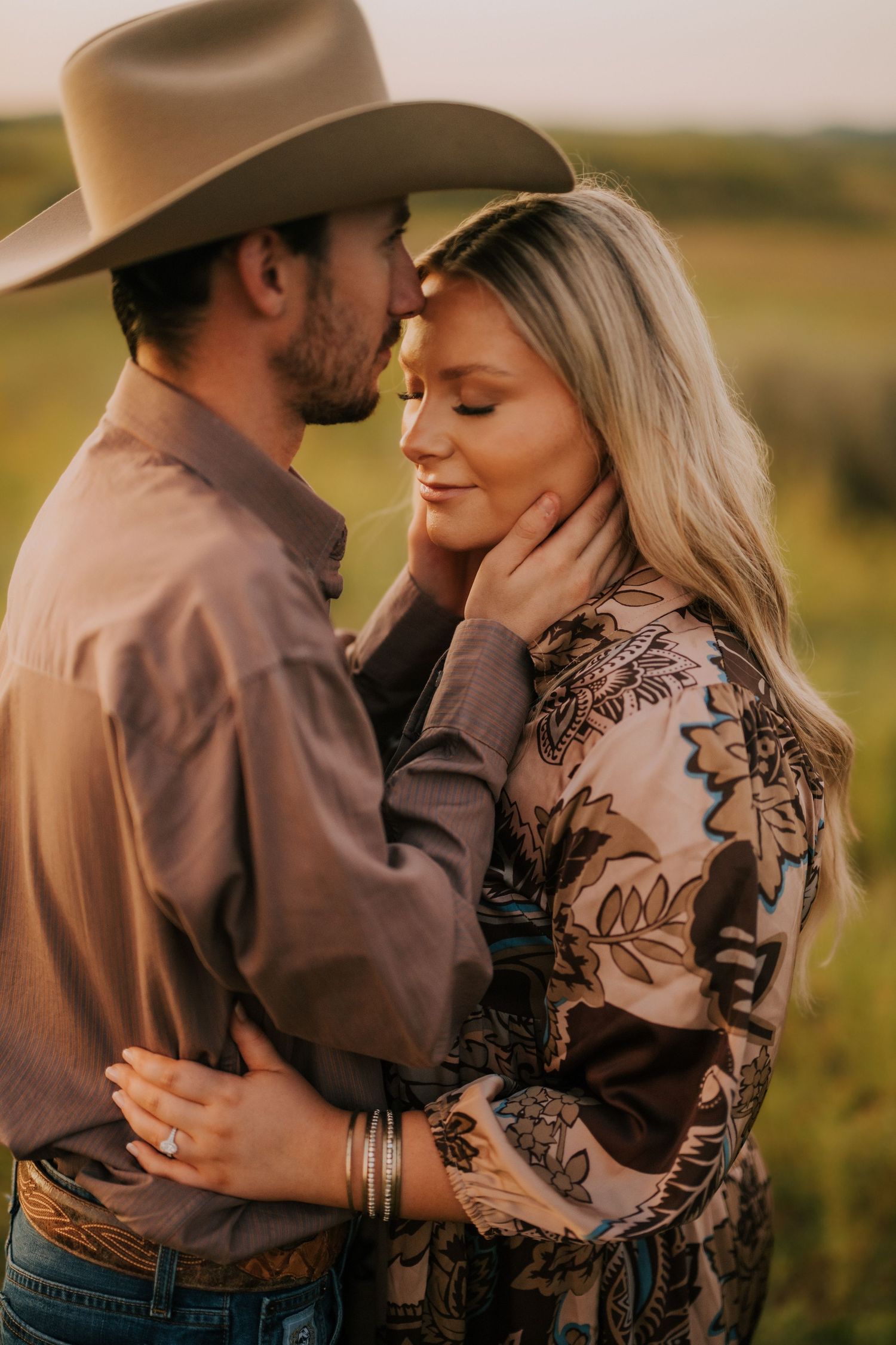 Romantic western-themed couple embraces in field wearing brown and patterned outfits.