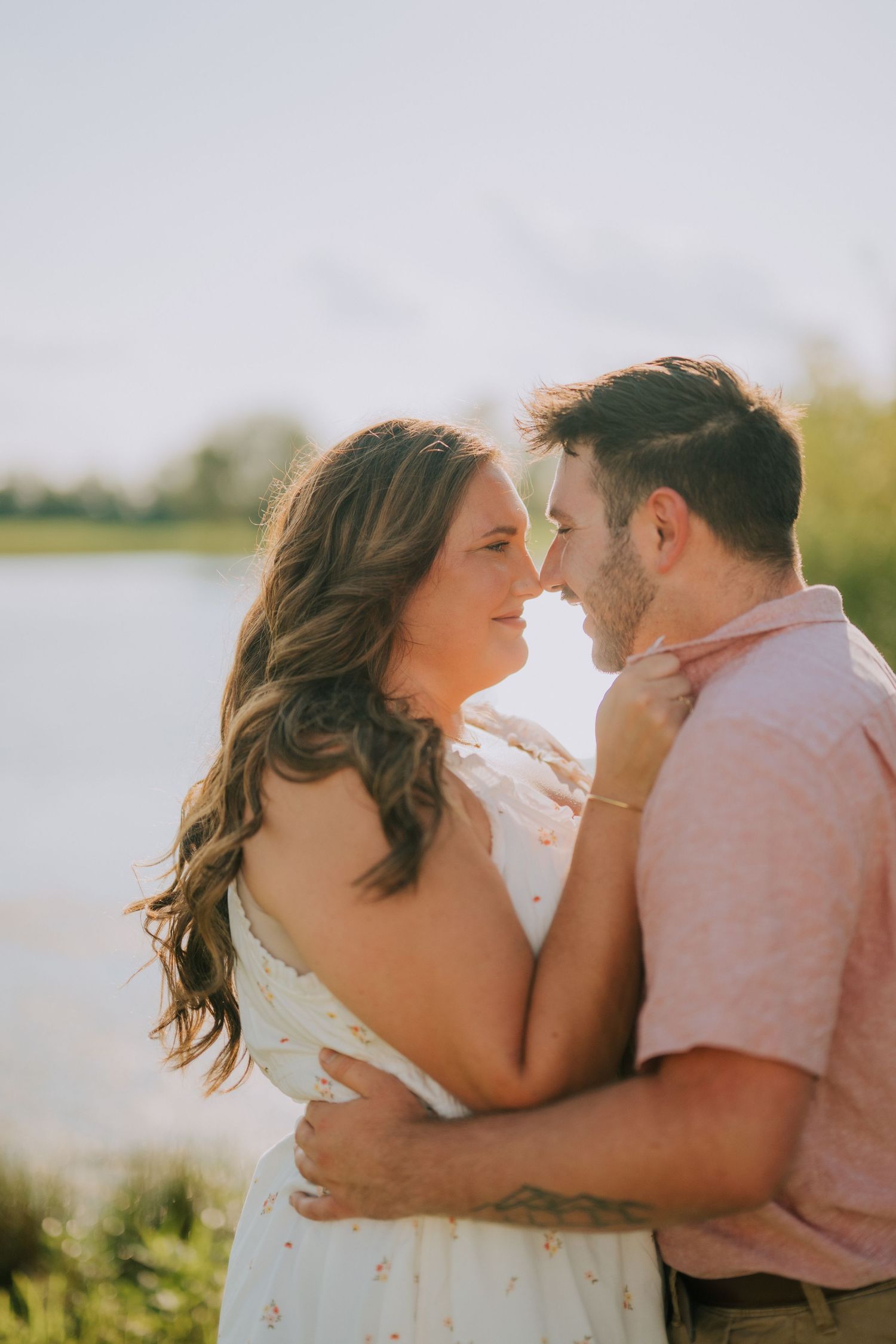 Romantic couple in white dress and pink shirt share intimate moments by the water during golden hour.