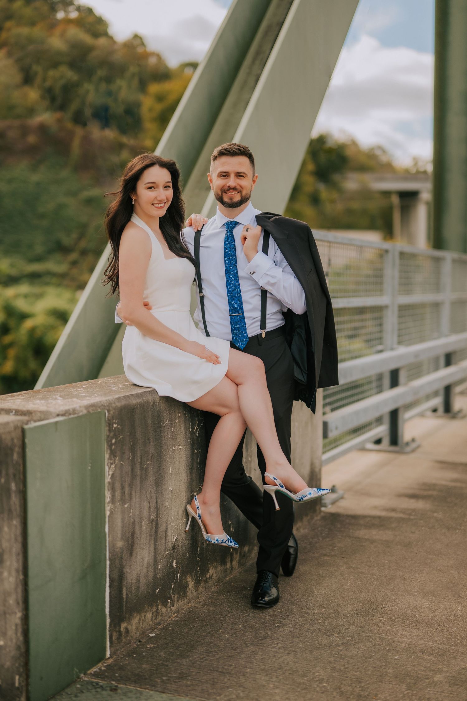 Couple sitting together on a bridge railing in formal attire.