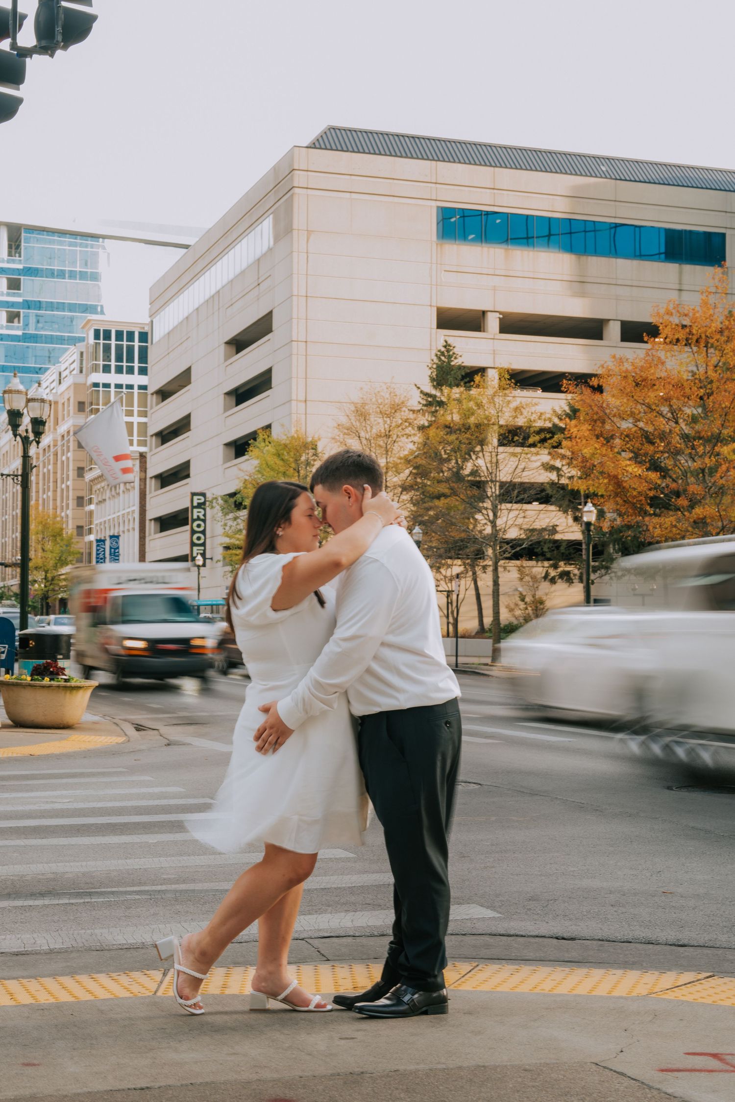 A romantic urban photoshoot with modern buildings and autumn trees in the background.