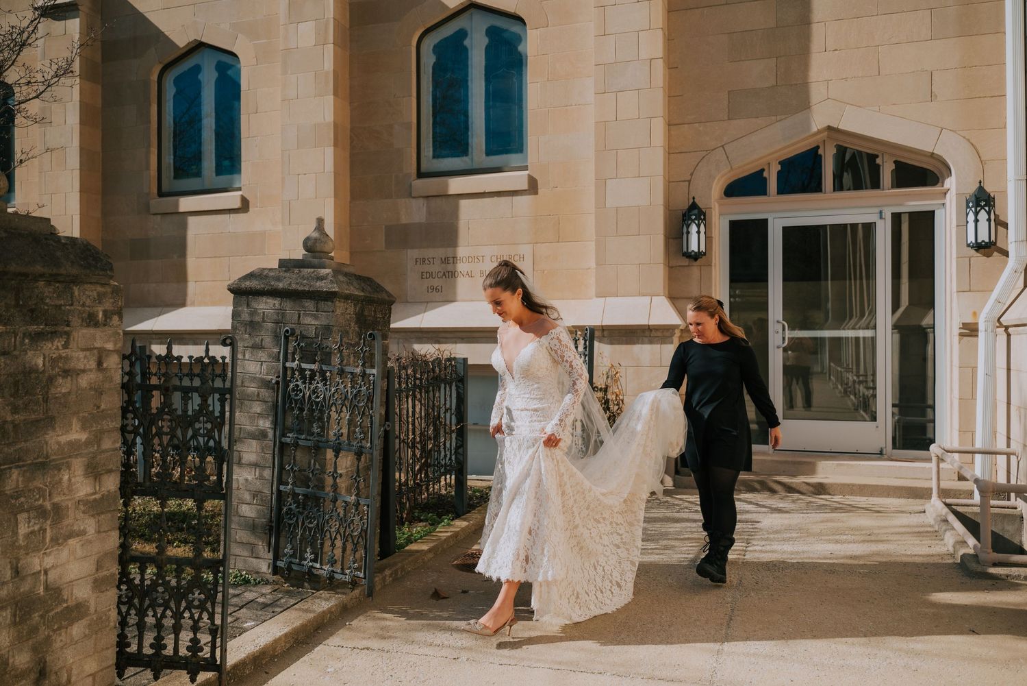 A couple in formal attire walks together through an elegant stone archway with wrought iron gates at a historic venue.