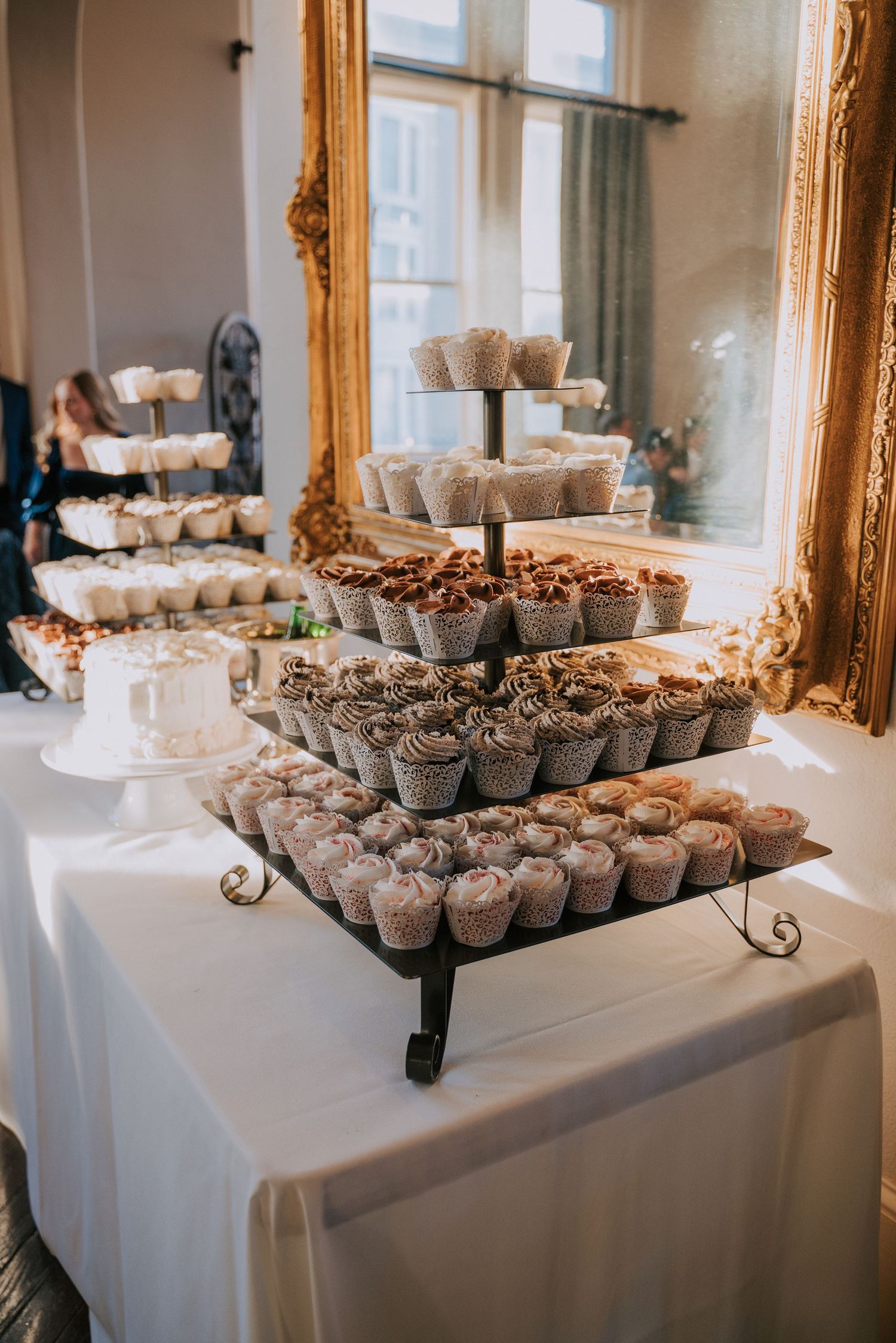Elegant tiered display of cupcakes arranged on white tablecloth with ornate gold mirror frame in background.