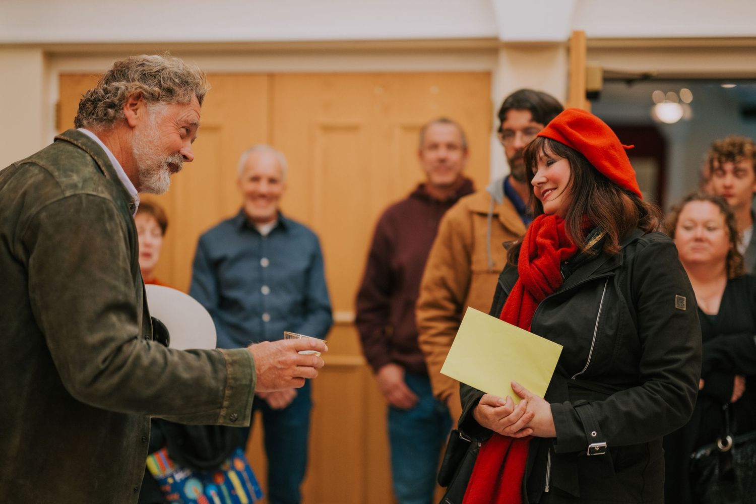 People gathered in an indoor meeting space as a person in a green jacket speaks with someone wearing a red scarf.