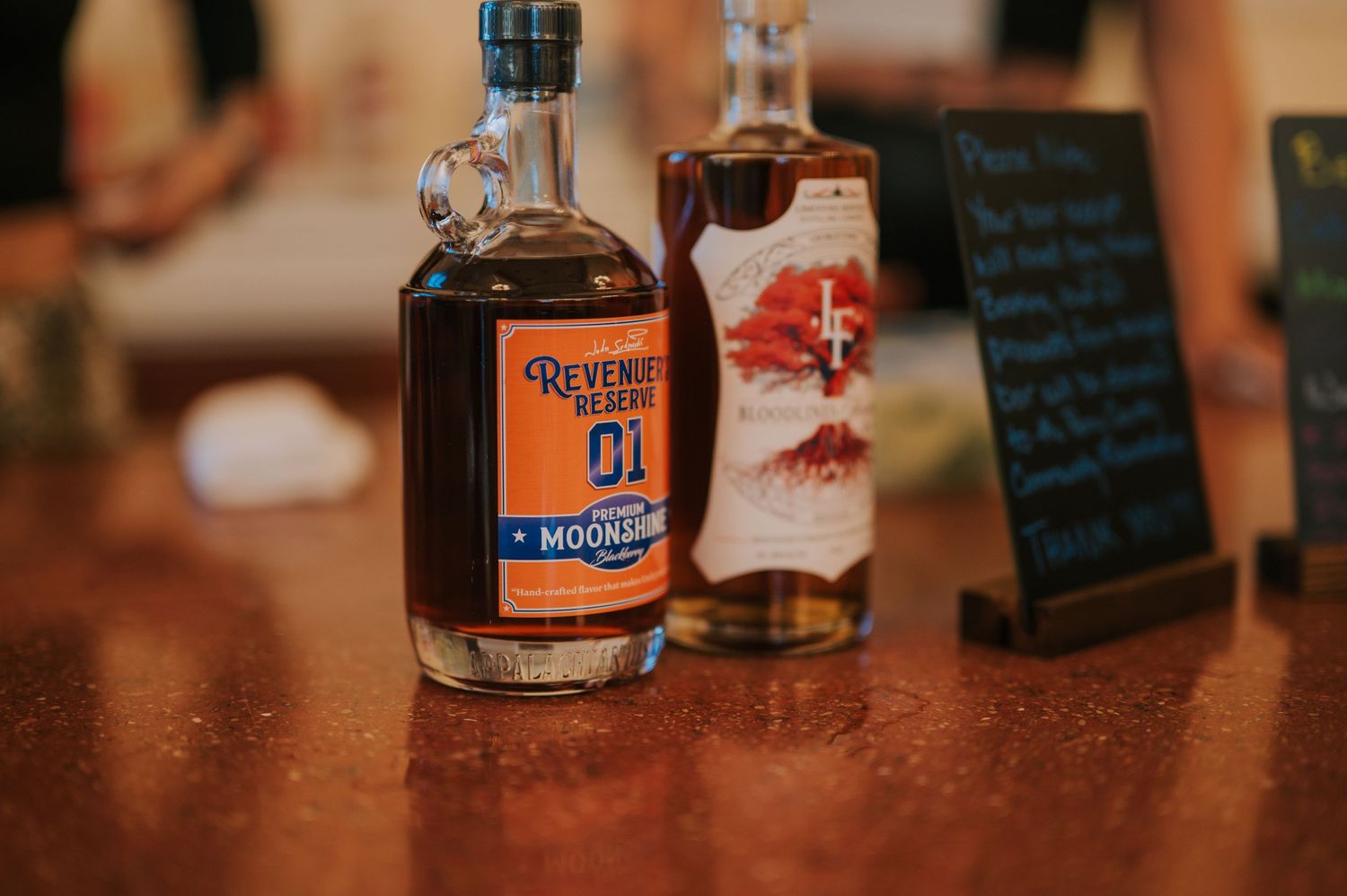 Two bottles of bourbon whiskey stand on a wooden bar counter with a small chalkboard menu in the background.