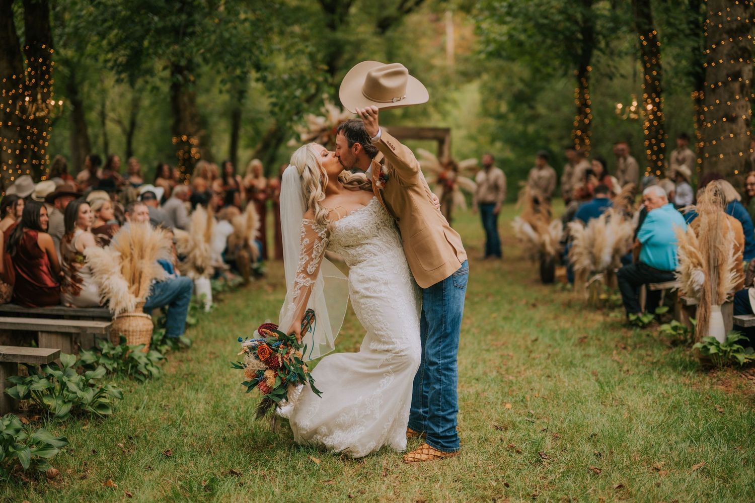 Bride and groom share a romantic kiss under a cowboy hat at an outdoor rustic woodland wedding ceremony.