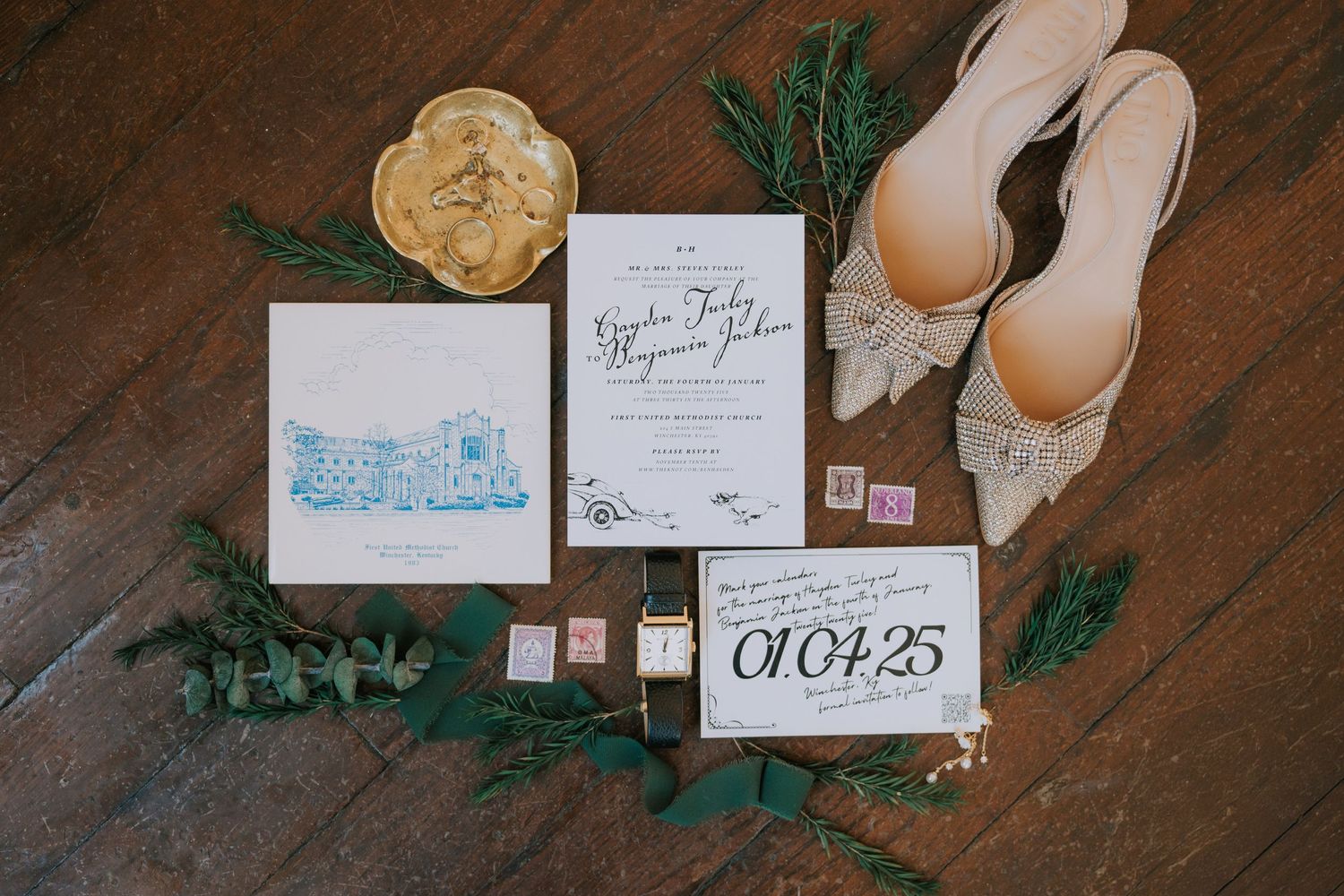 Wedding details flat lay with invitation suite, white shoes, and greenery on dark wooden floor.