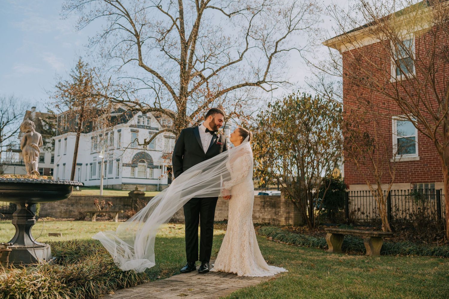 A couple shares a romantic moment in a garden setting as the bride's long veil flows gracefully in the autumn breeze.