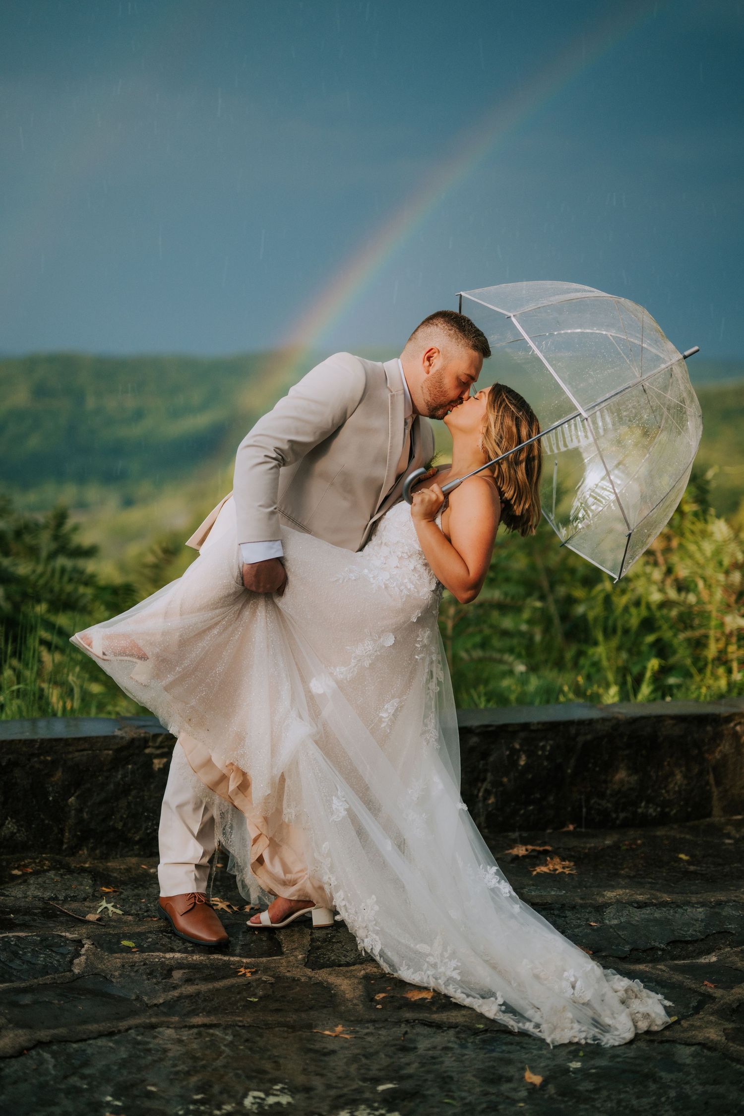 A romantic wedding photoshoot during sunset with a rainbow and clear umbrella on a hilltop.