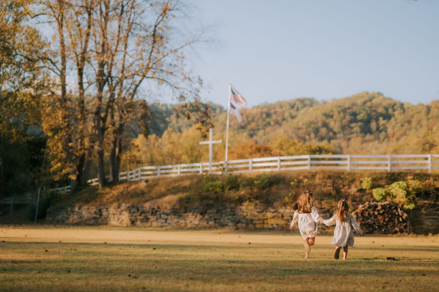 Panoramic view of wedding party gathered in rural field with mountains and white fence.