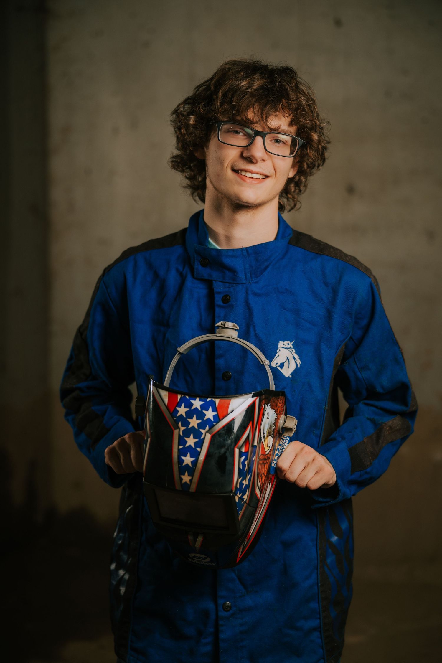 Student displays welding equipment while wearing protective blue workshop gear.