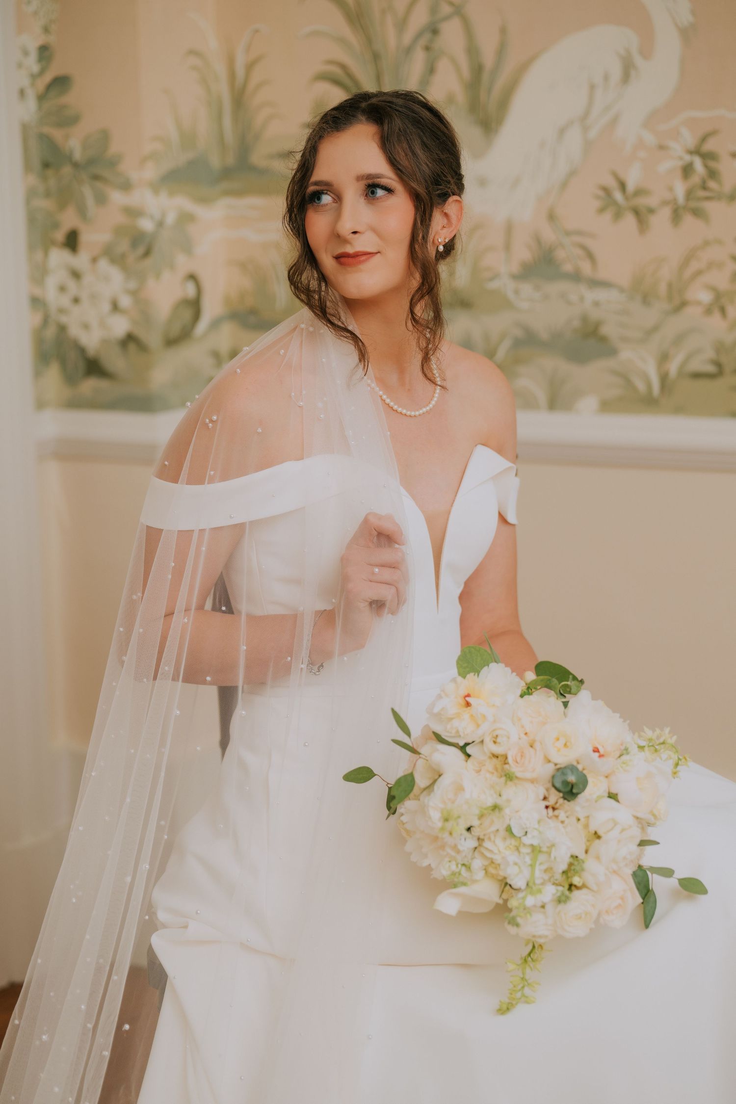 Bride in off-shoulder wedding gown holding white floral bouquet against floral wallpaper backdrop.