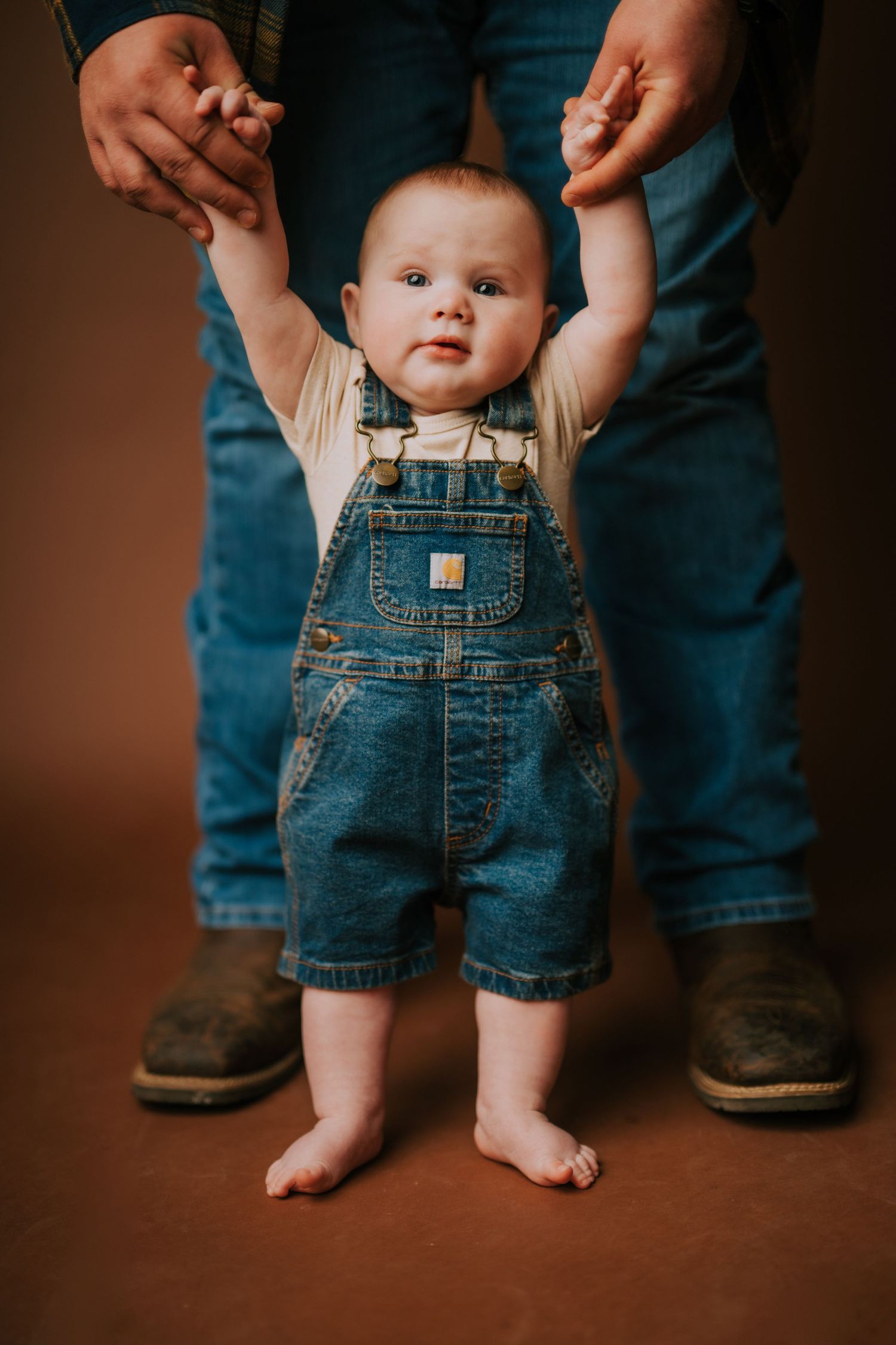 Small baby wearing denim overalls stands between adult legs against brown background.