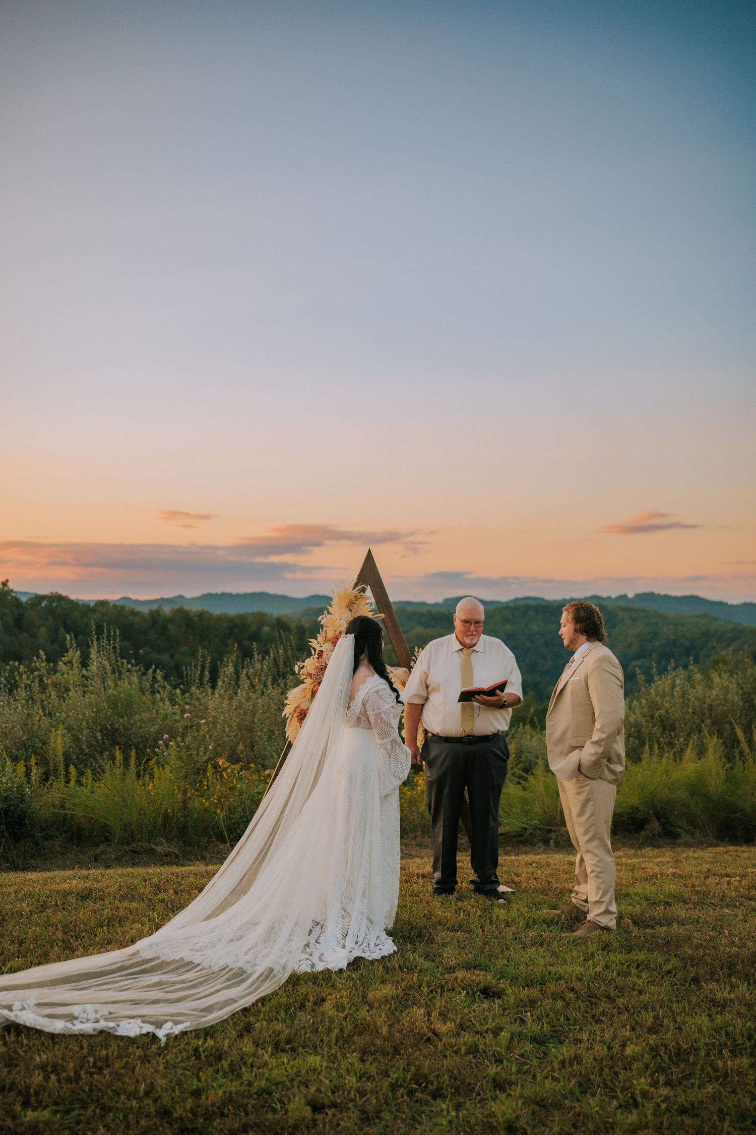 A wedding party stands on a hilltop at sunset with mountains in the background and flowing white dresses in view.