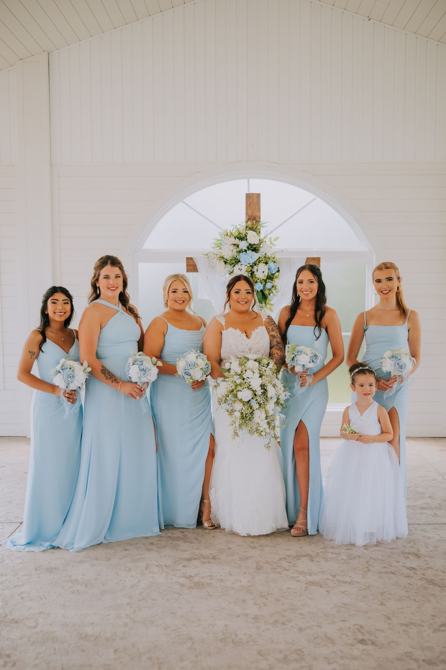 Bridesmaids in light blue dresses pose with the bride under a white archway.