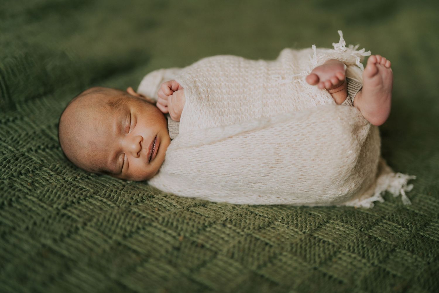 A sleeping newborn wrapped in white knit blanket rests on green textured fabric.