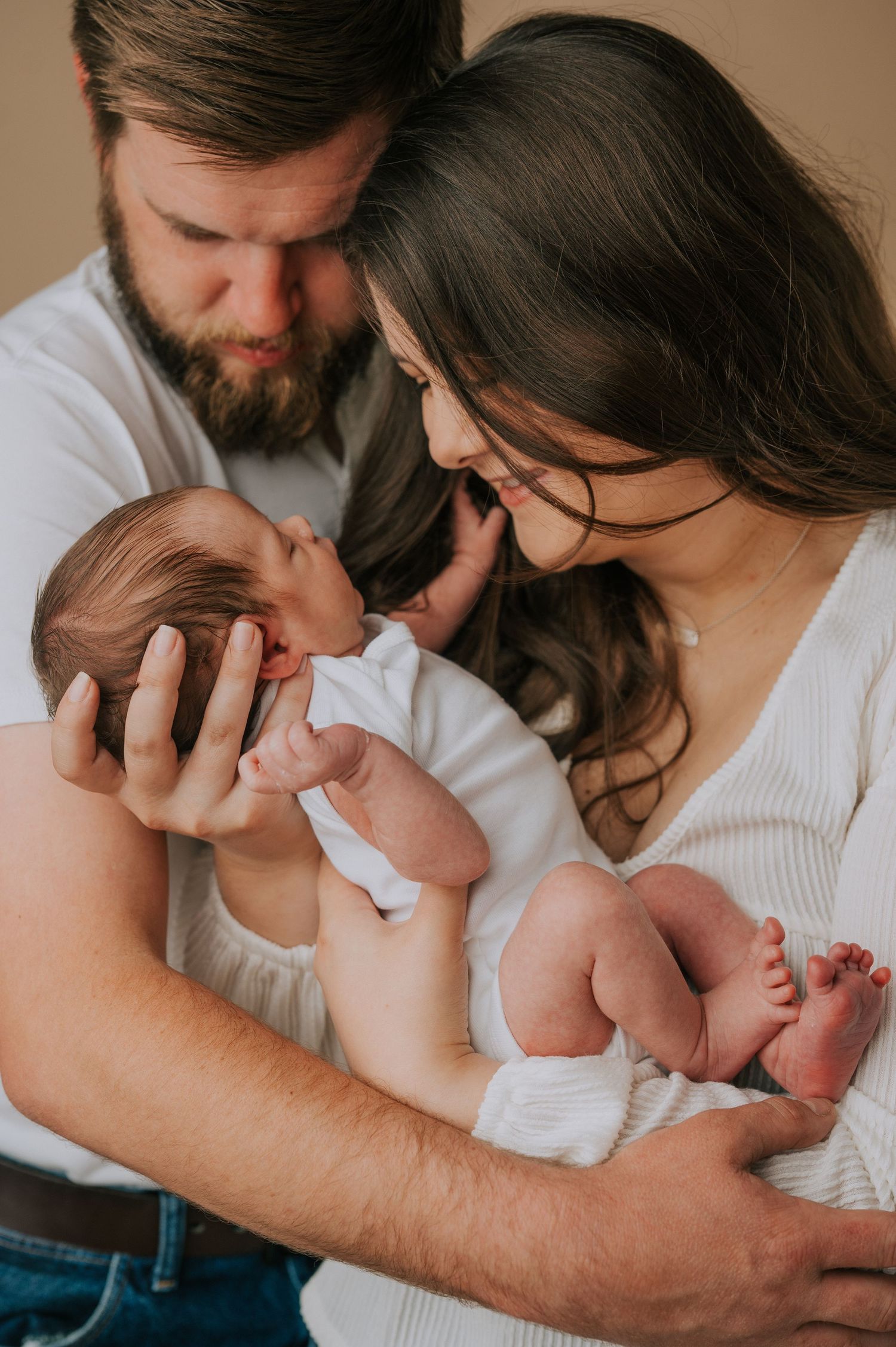 Parents sharing intimate moment with newborn baby against neutral beige background.