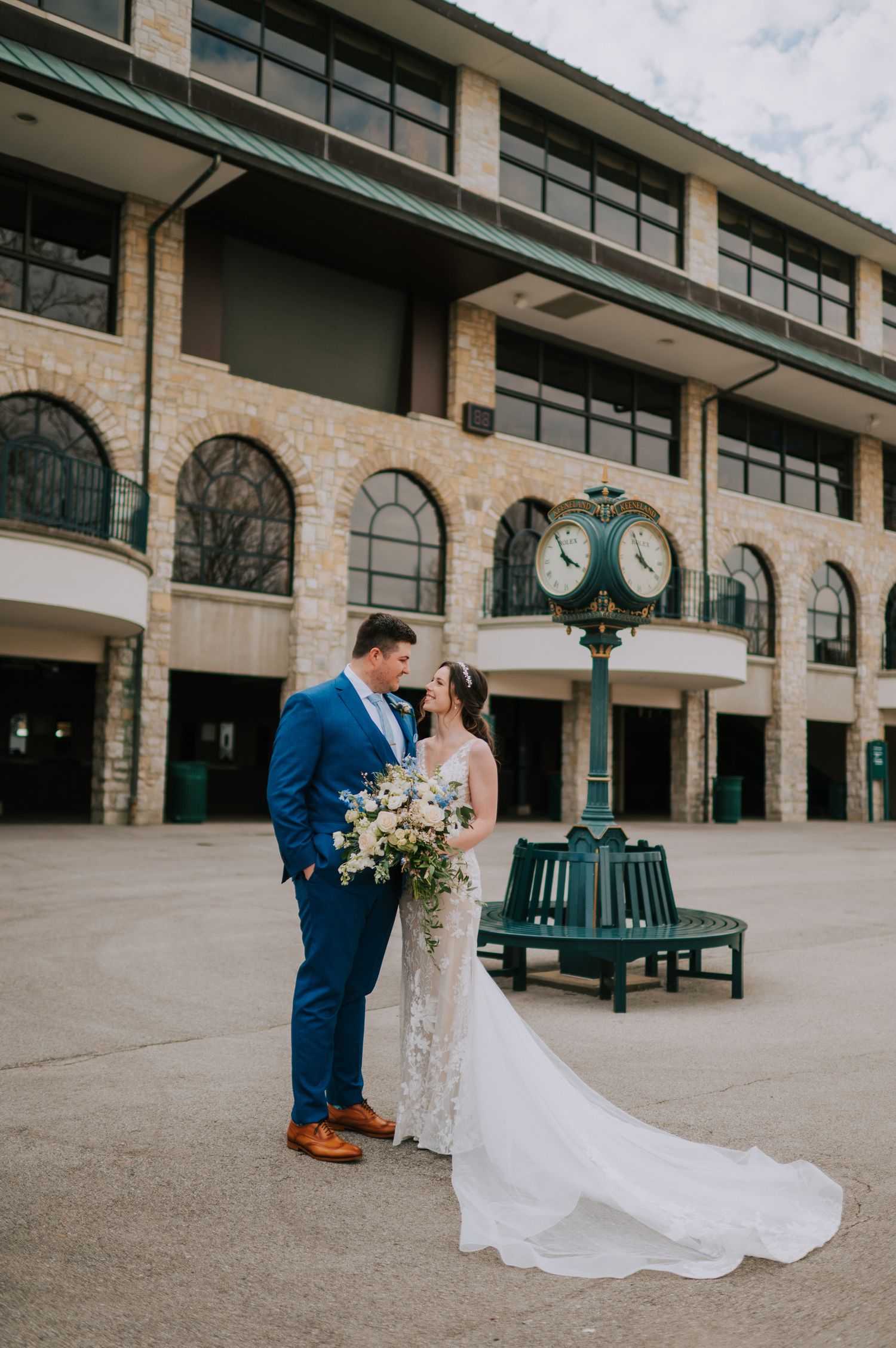 Wedding couple in blue suit and white dress embracing outside stone building.