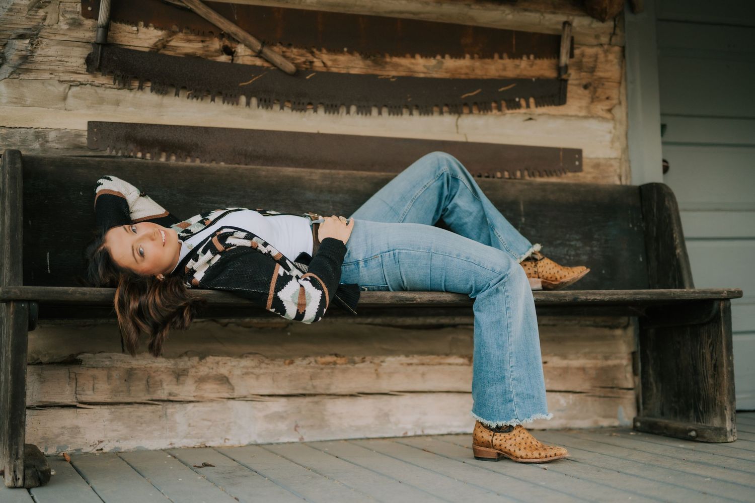 Person lying casually on wooden porch steps wearing jeans and boots in artistic portrait composition.
