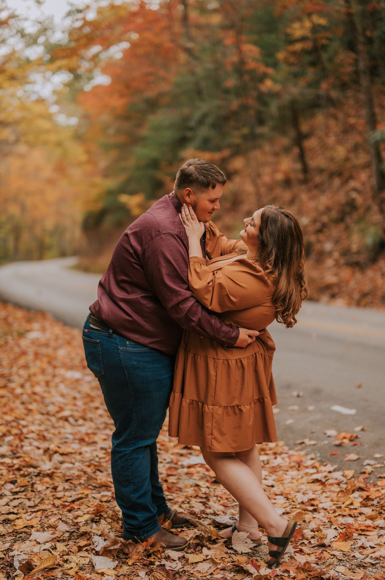 A romantic autumn photoshoot on a leaf-covered road with warm earth-toned outfits and fall foliage in the background.