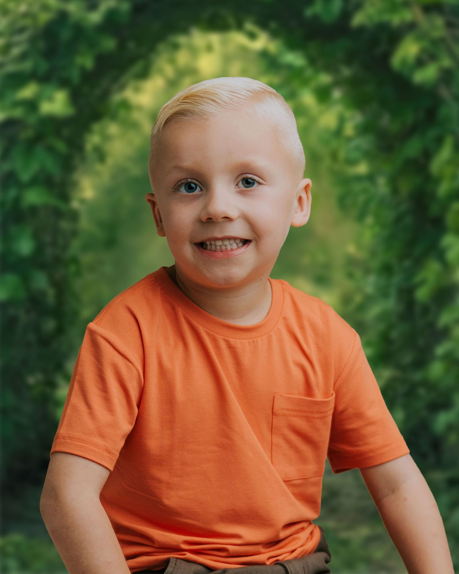 Smiling blonde boy in orange t-shirt posing outdoors with lush green arched foliage background.
