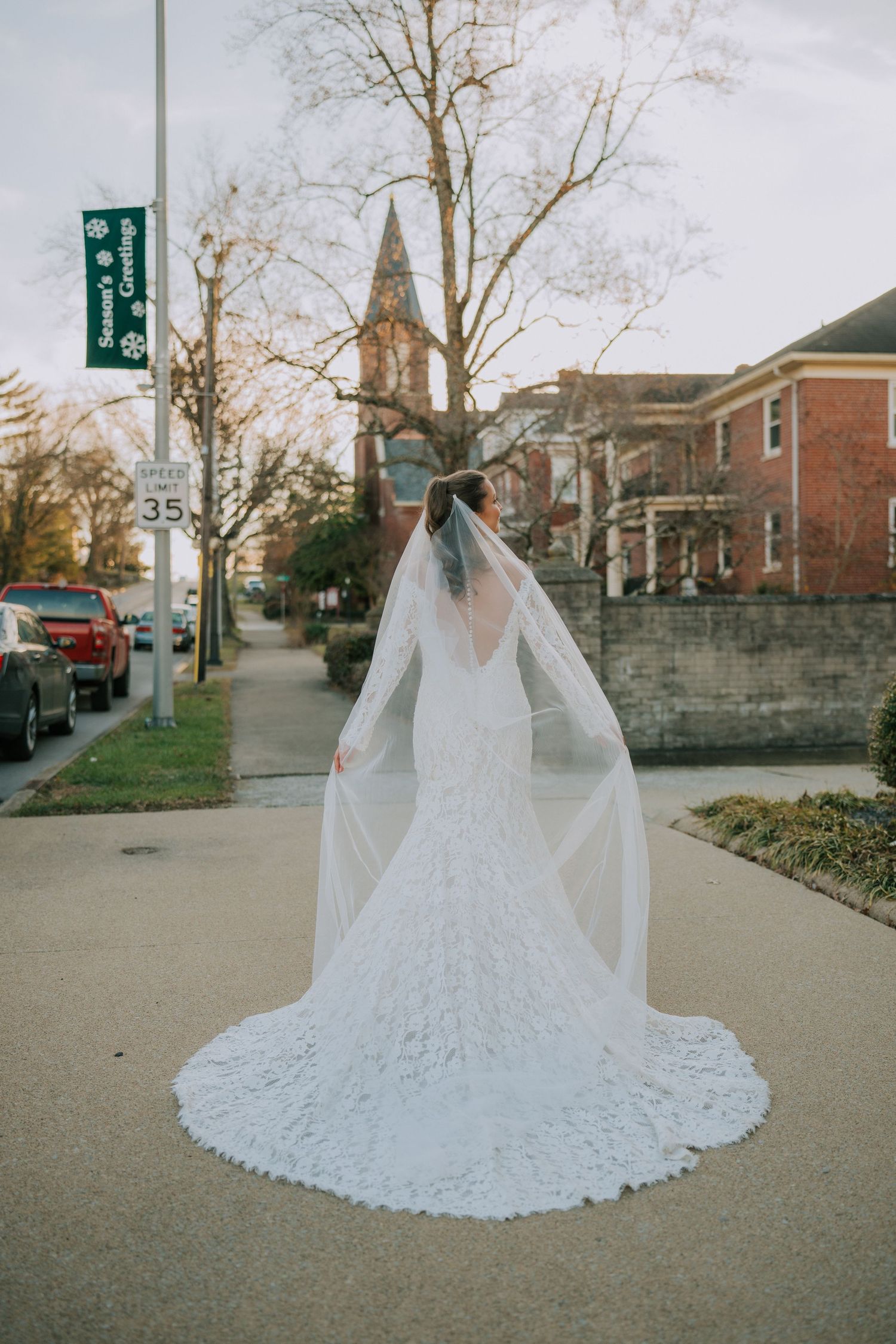 A detailed lace wedding dress with flowing veil photographed on a city sidewalk with a church steeple in the background.