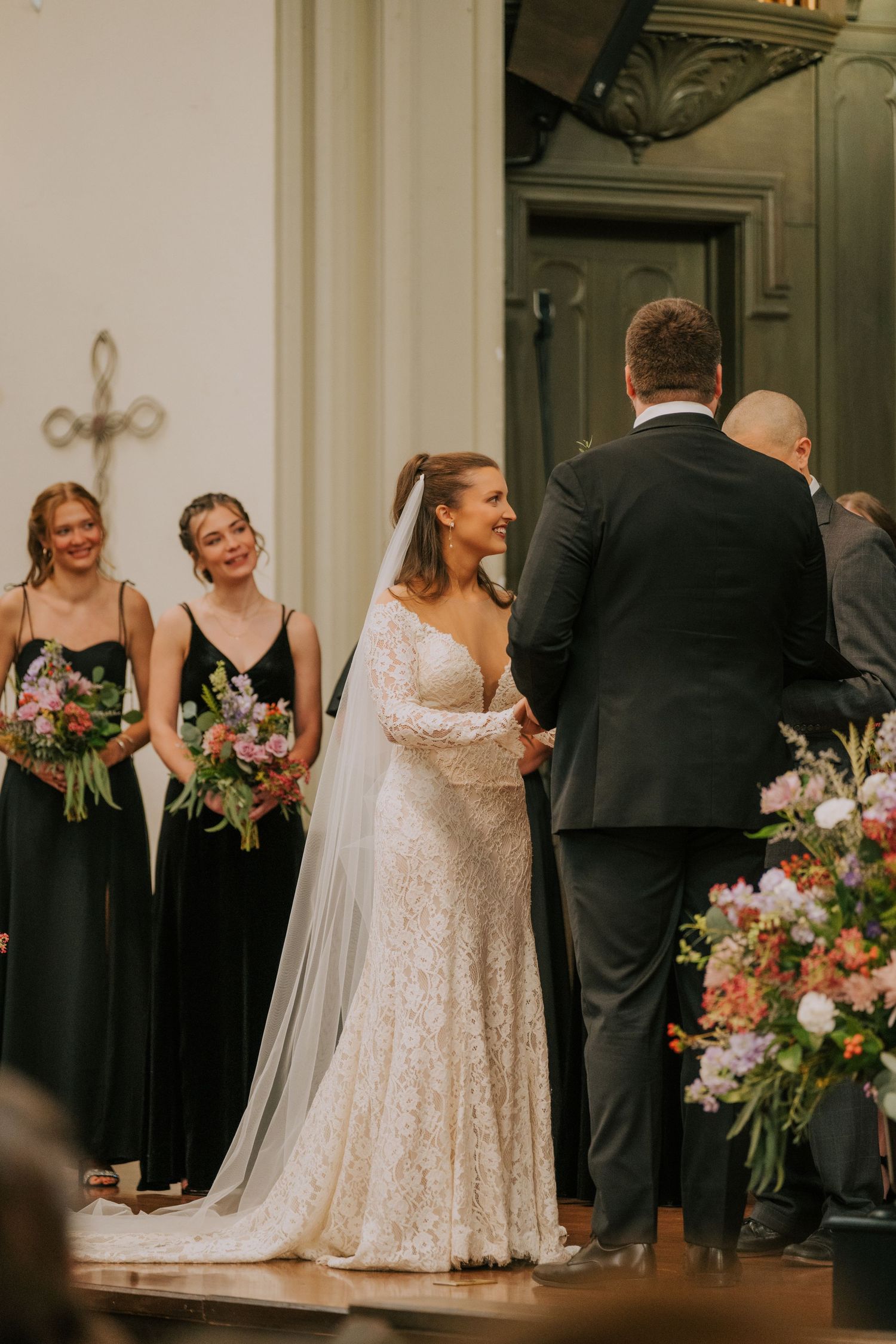 Beautiful wedding ceremony in progress with bridesmaids in black dresses admiring the couple at the altar with floral arrangements.