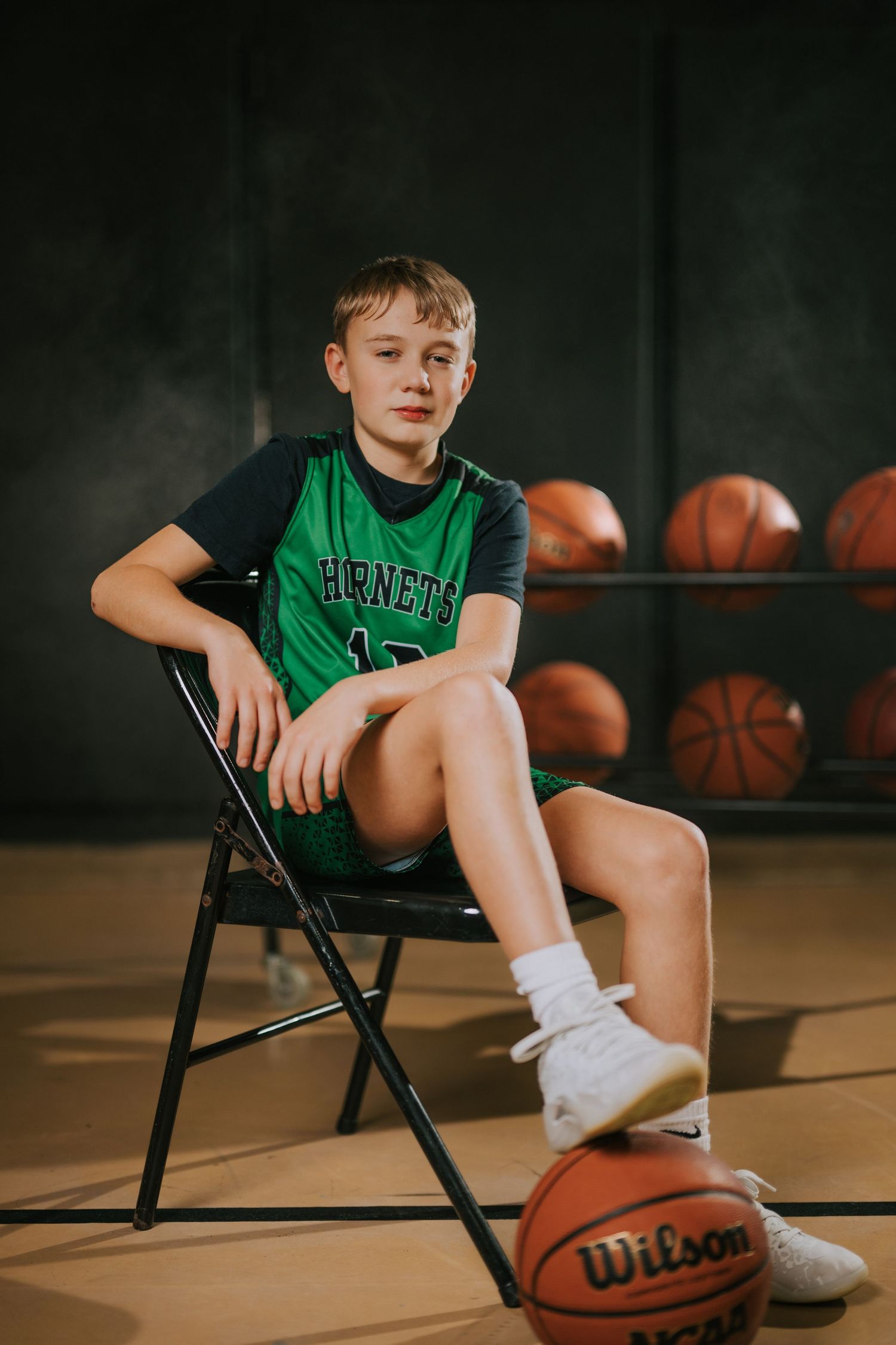 Young basketball player in green jersey sits on folding chair with basketballs visible in background.
