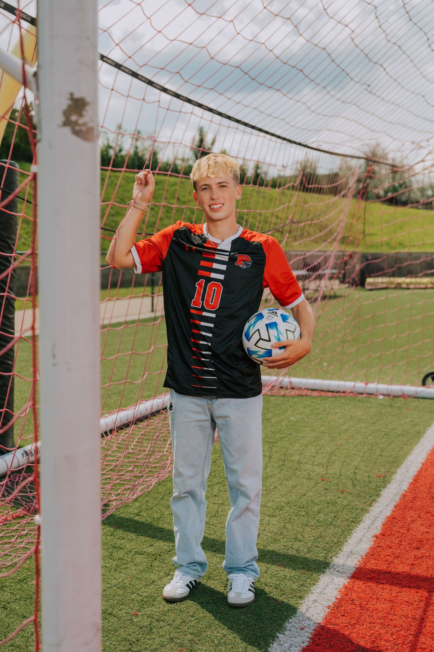 A person in jeans and a black shirt stands in a soccer goal with a ball on a sunny day.