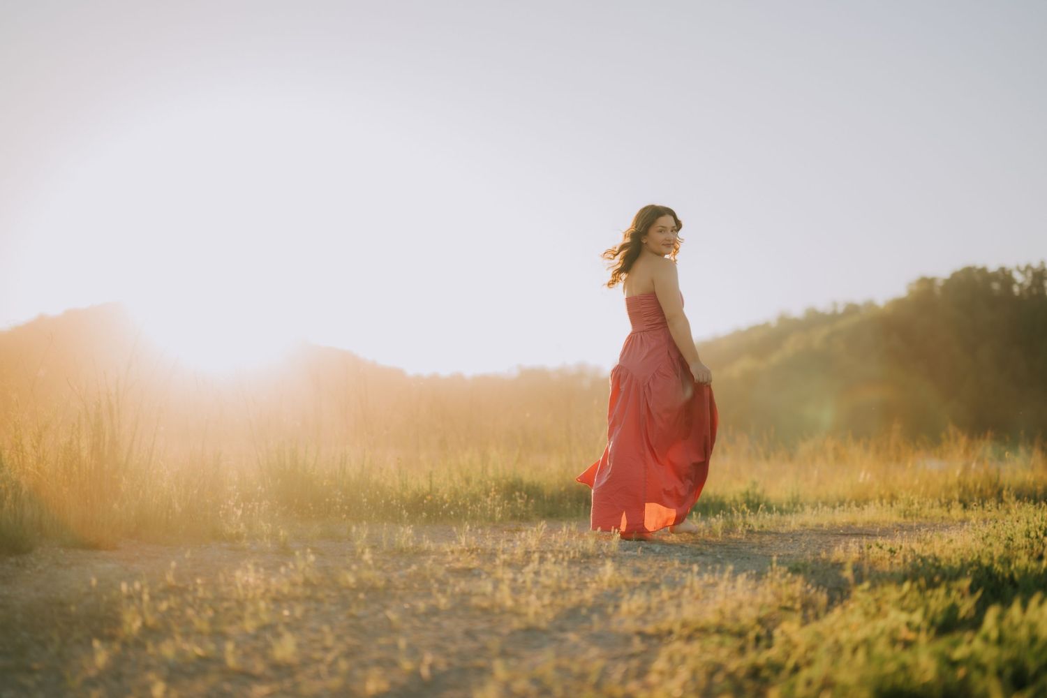 Woman in flowing red dress standing in golden sunlight on a dirt path surrounded by lush green meadows.