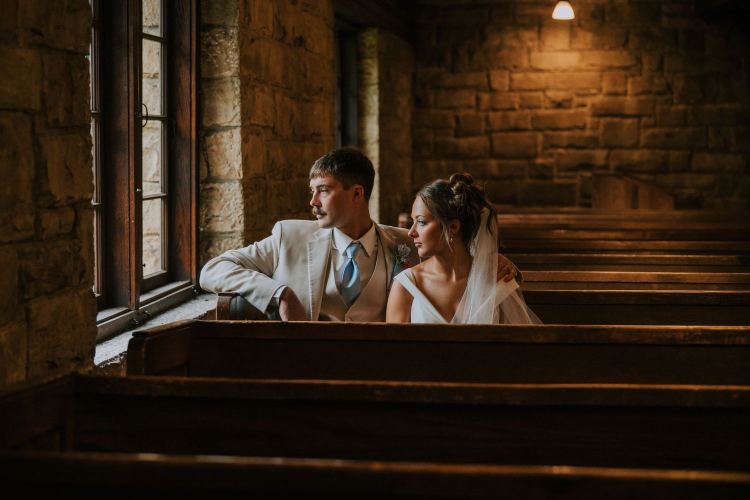 Romantic couple sits intimately in vintage church pews with exposed brick walls and warm lighting creating moody ambiance.