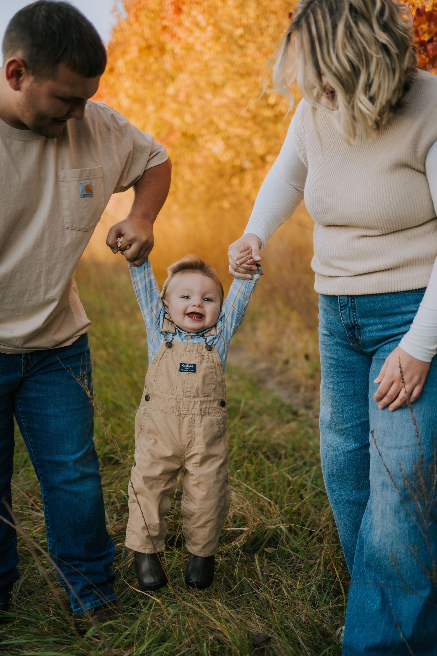 A toddler in khaki shorts walks through grass with adults in casual clothing during a sunny autumn day.