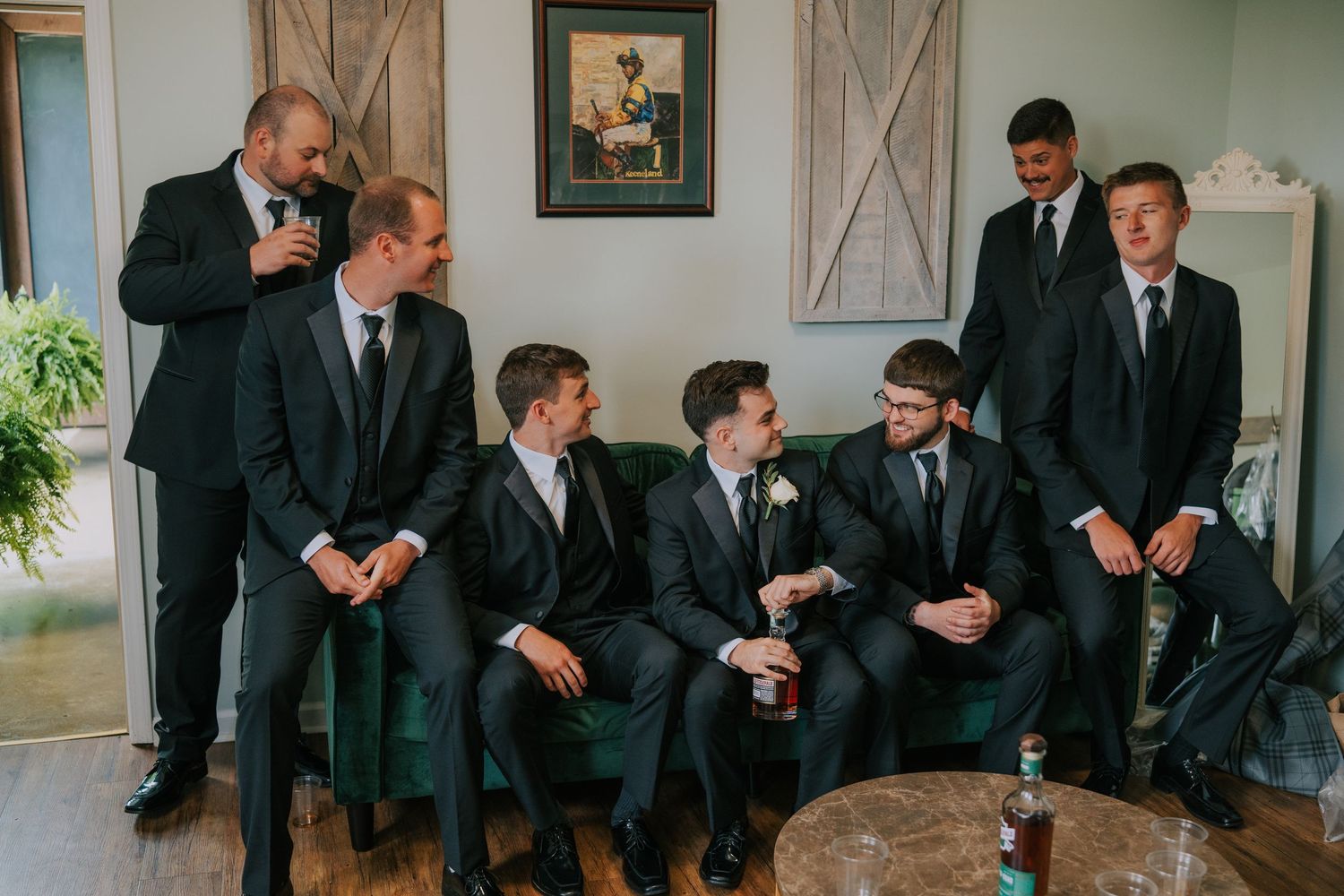 Group of groomsmen in black suits gathered together in a rustic indoor setting.