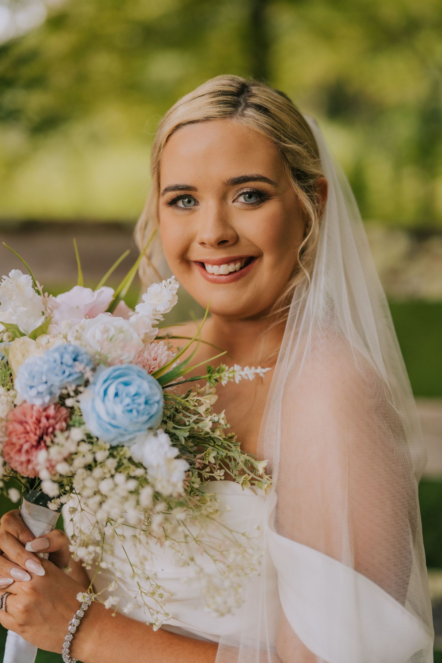 A bride holds a pastel bouquet of blue and pink flowers while wearing a white dress and veil.