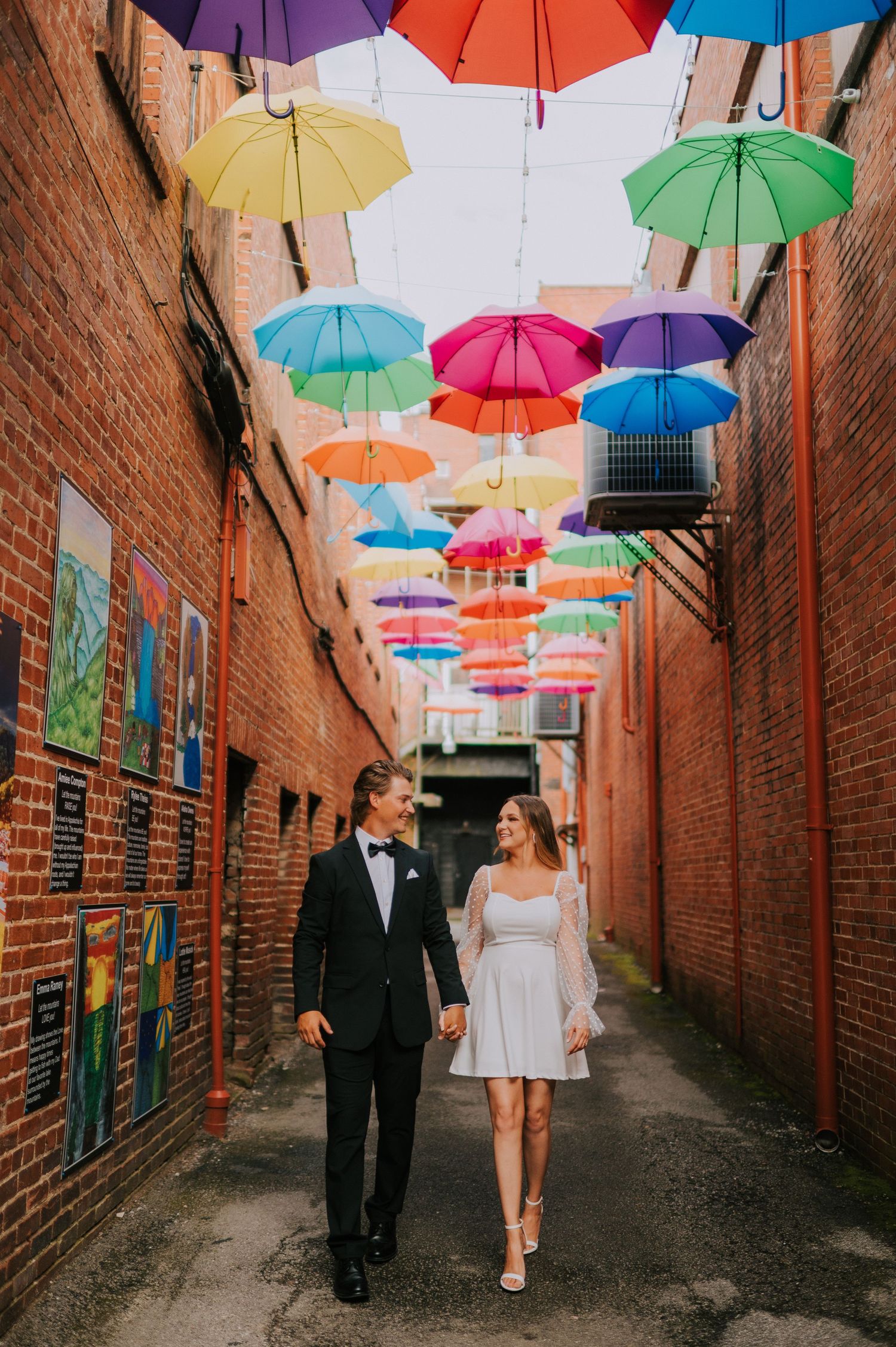 A couple walks beneath colorful suspended umbrellas in a charming brick alleyway.