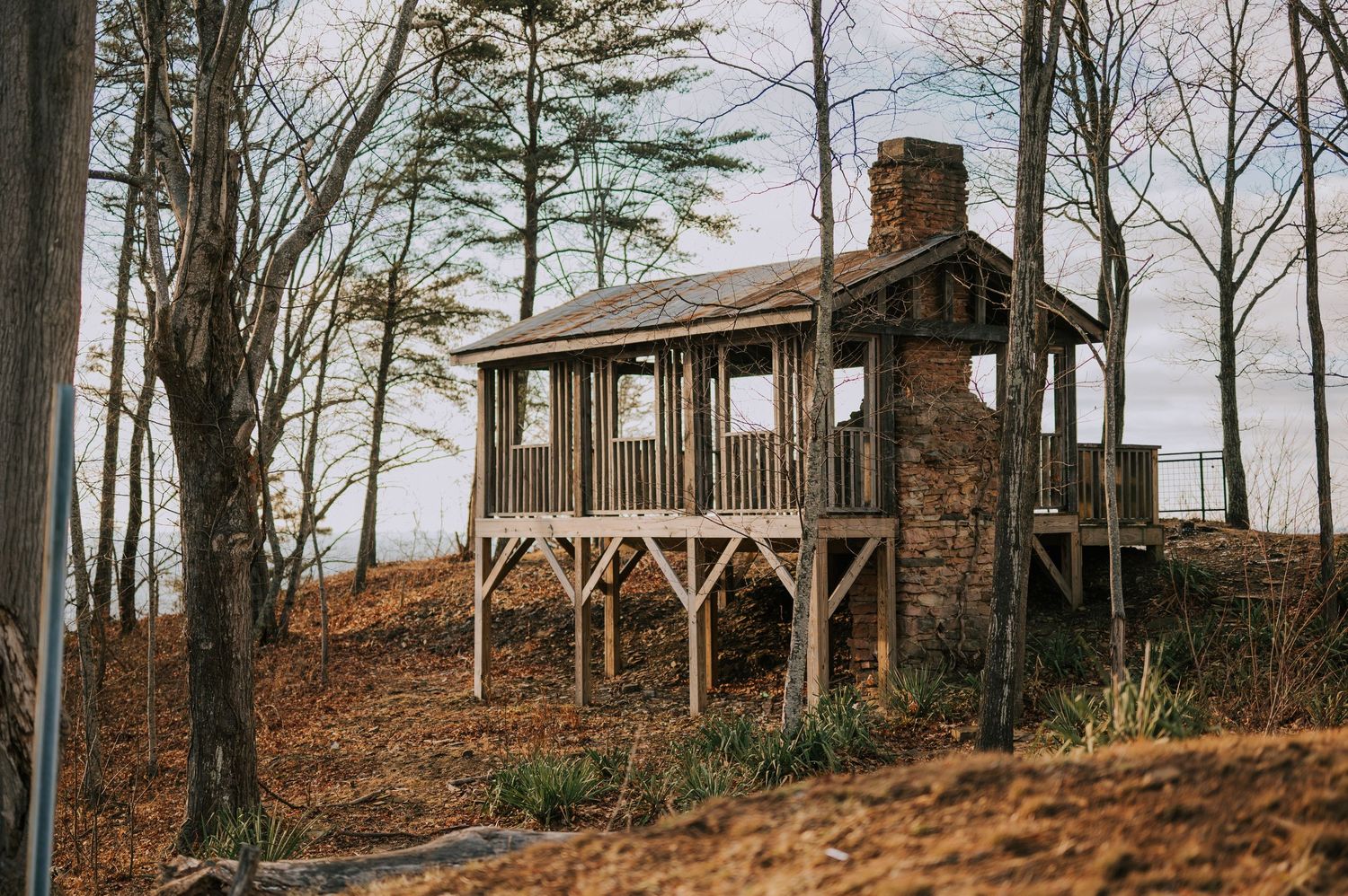 A rustic stone and wood cabin sits atop a hill surrounded by bare trees in winter.