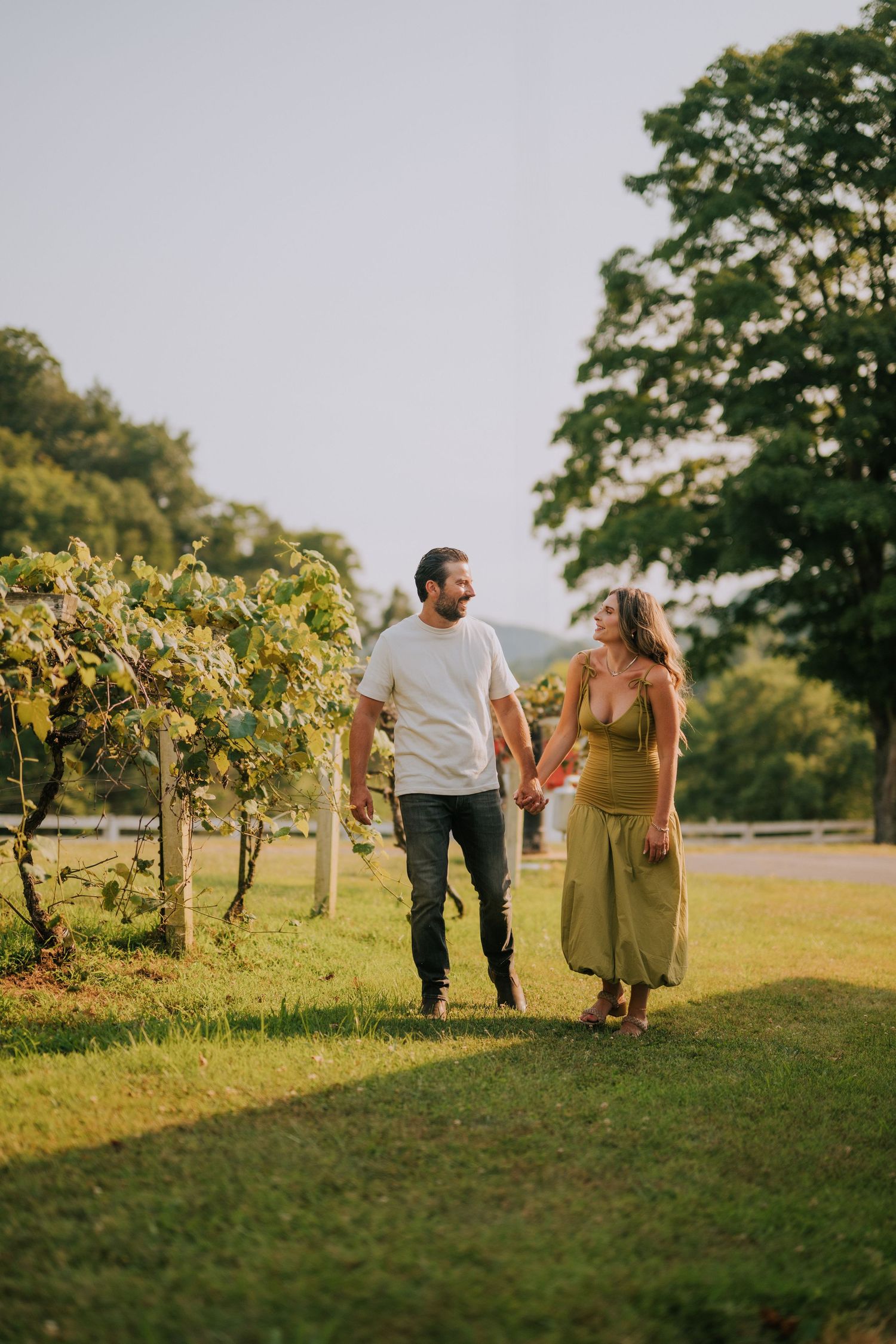 Couple strolling through a vineyard at sunset with golden light filtering through the trees.