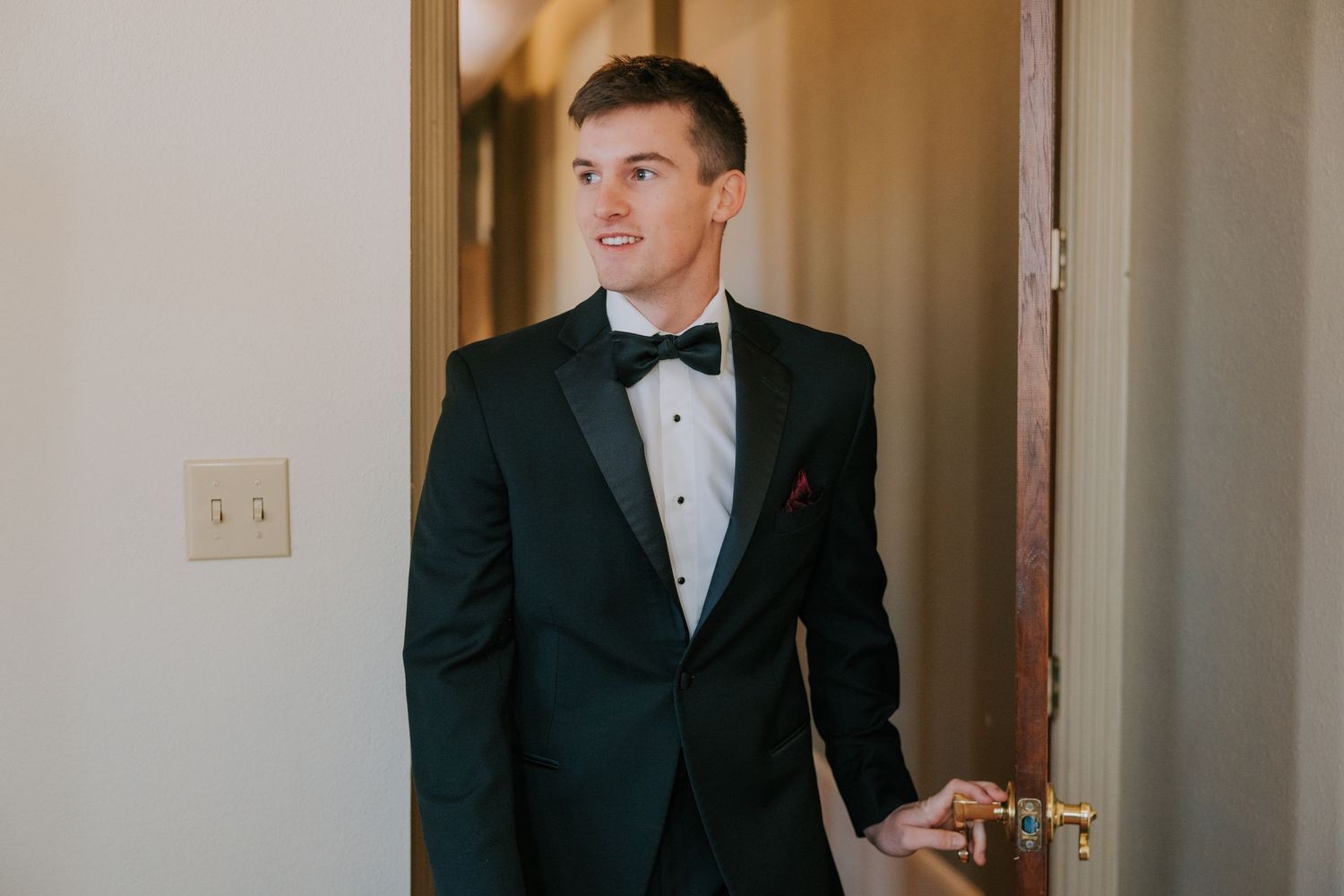 Person wearing a classic black tuxedo and bow tie stands in a hallway near a doorway.