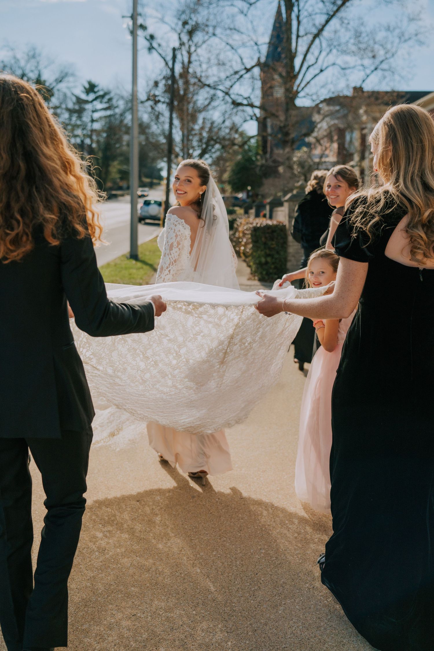 Bridesmaids in black dresses help adjust a flowing white wedding gown train outdoors near a church.