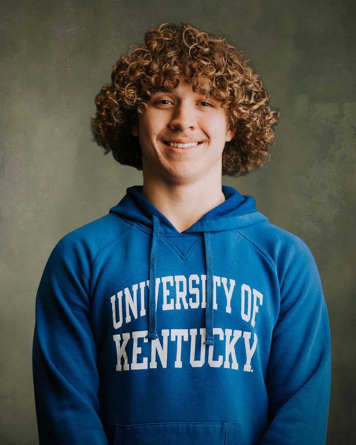 Smiling young man with curly hair wearing a blue University of Kentucky hoodie sweatshirt.