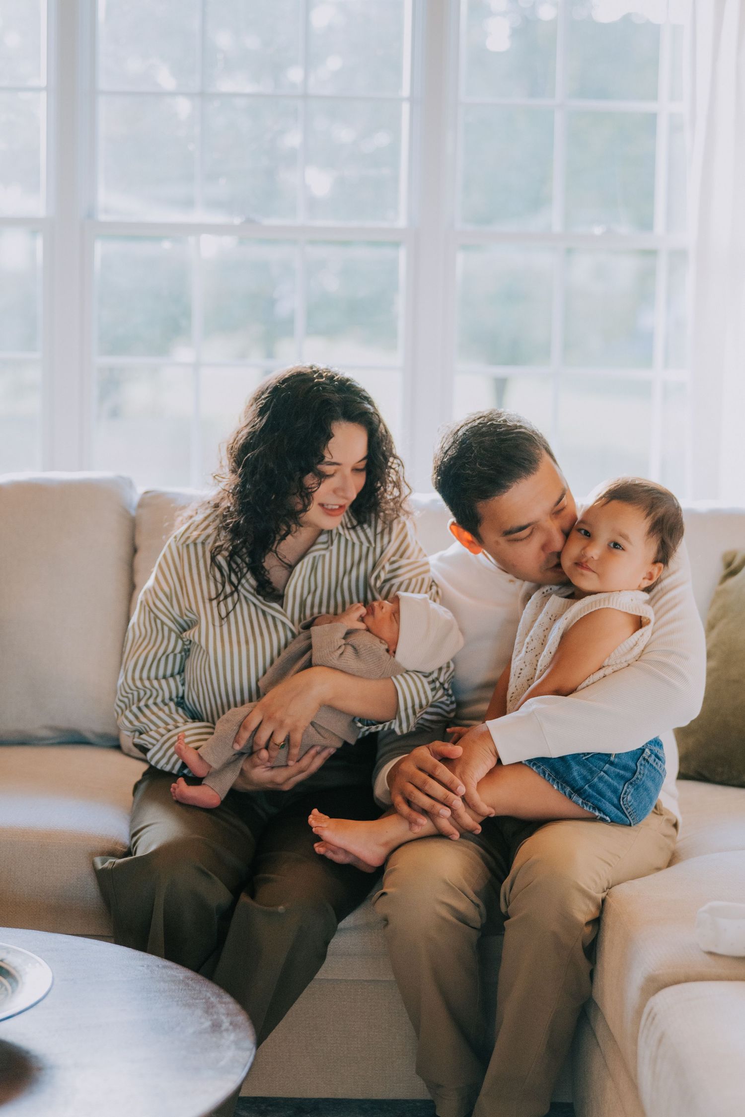 A tender family moment on a couch as parents snuggle with their baby in a bright, sunlit living room.
