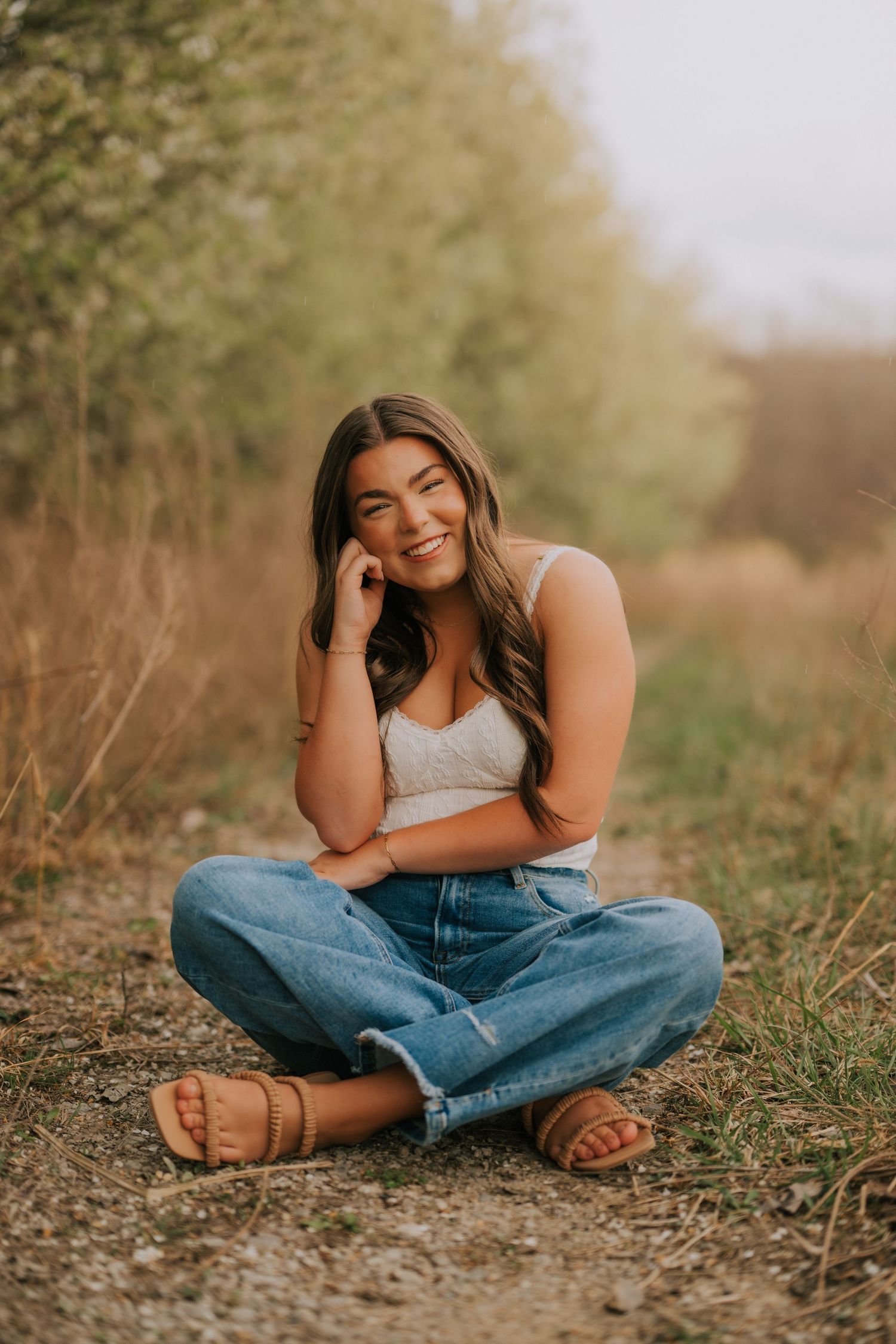 Casual portrait of person in jeans sitting cross-legged on ground outdoors.