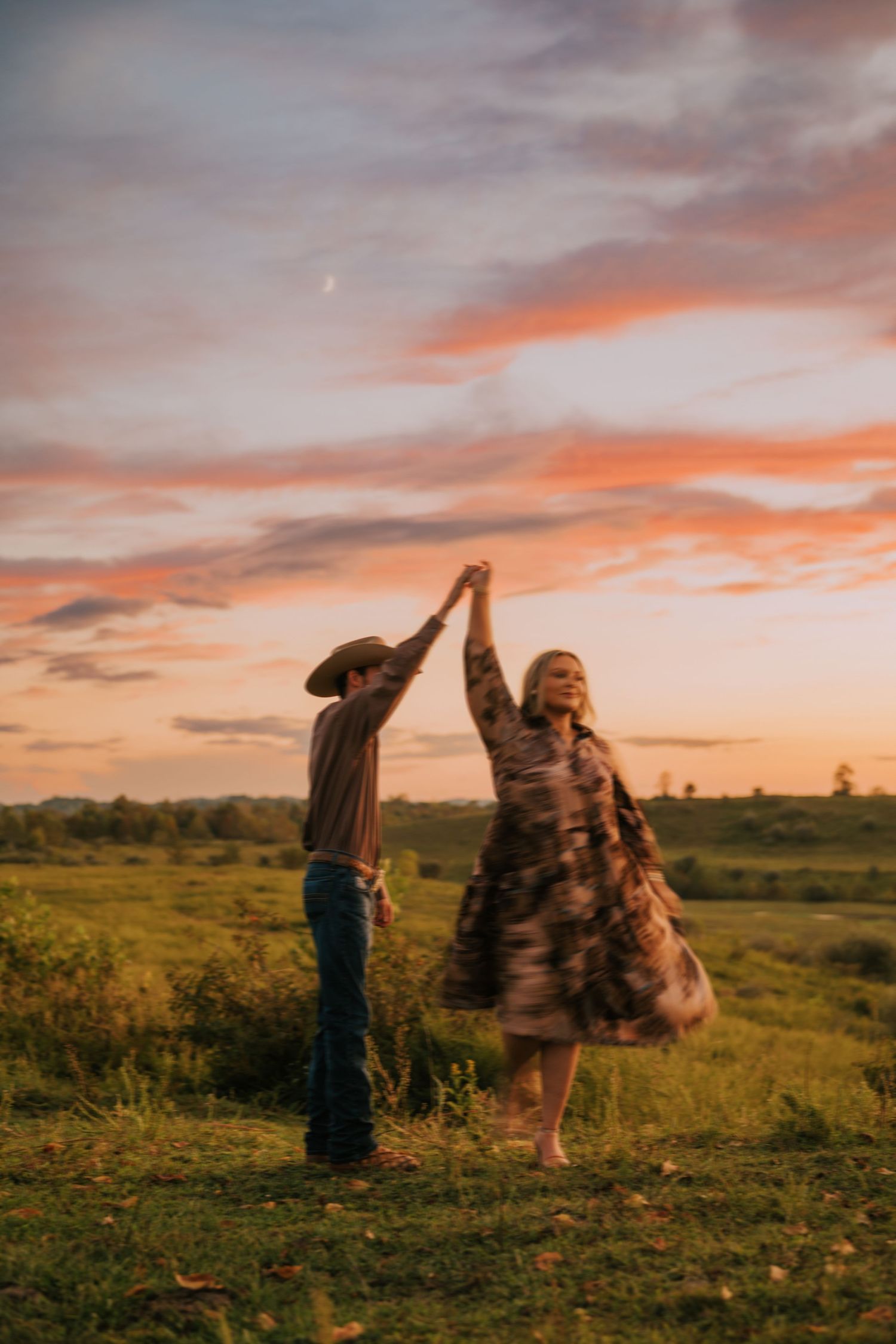 Couple dances in a field during golden hour with dramatic sunset clouds in the background.