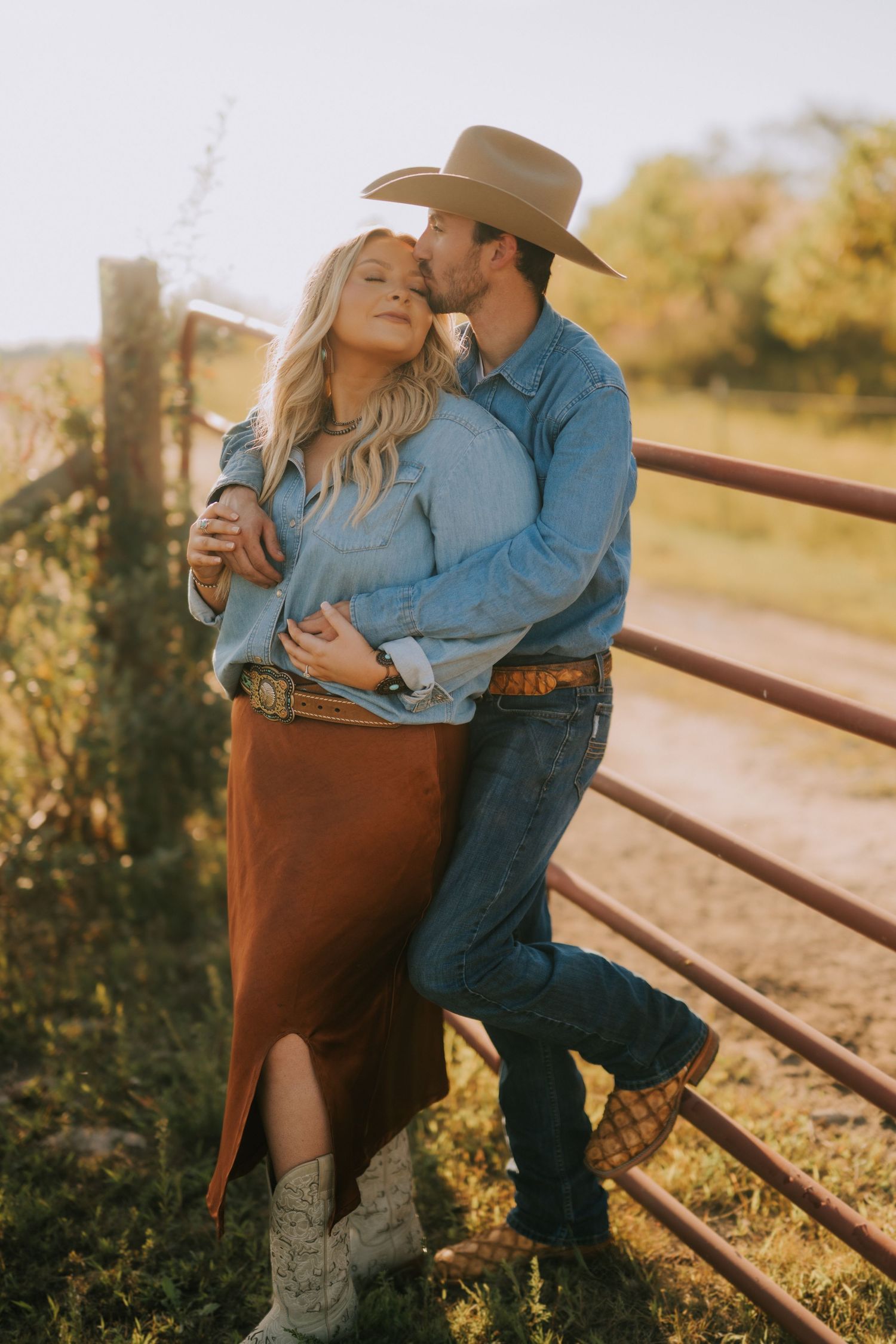 A romantic western-styled couple embraces against a rustic fence during golden hour in the countryside.