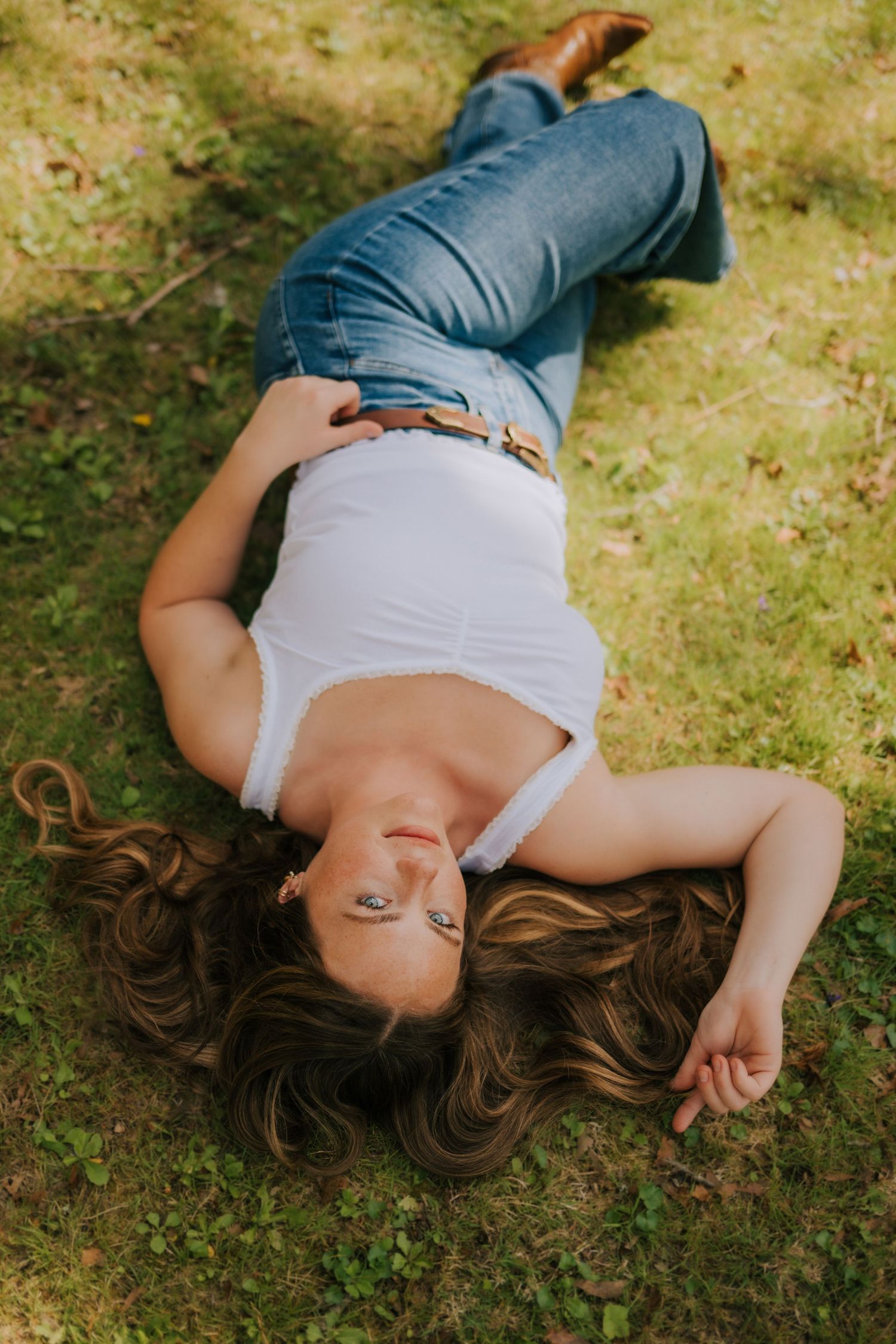 Woman in white tank top and jeans lying on green grass with long wavy hair spread around her.